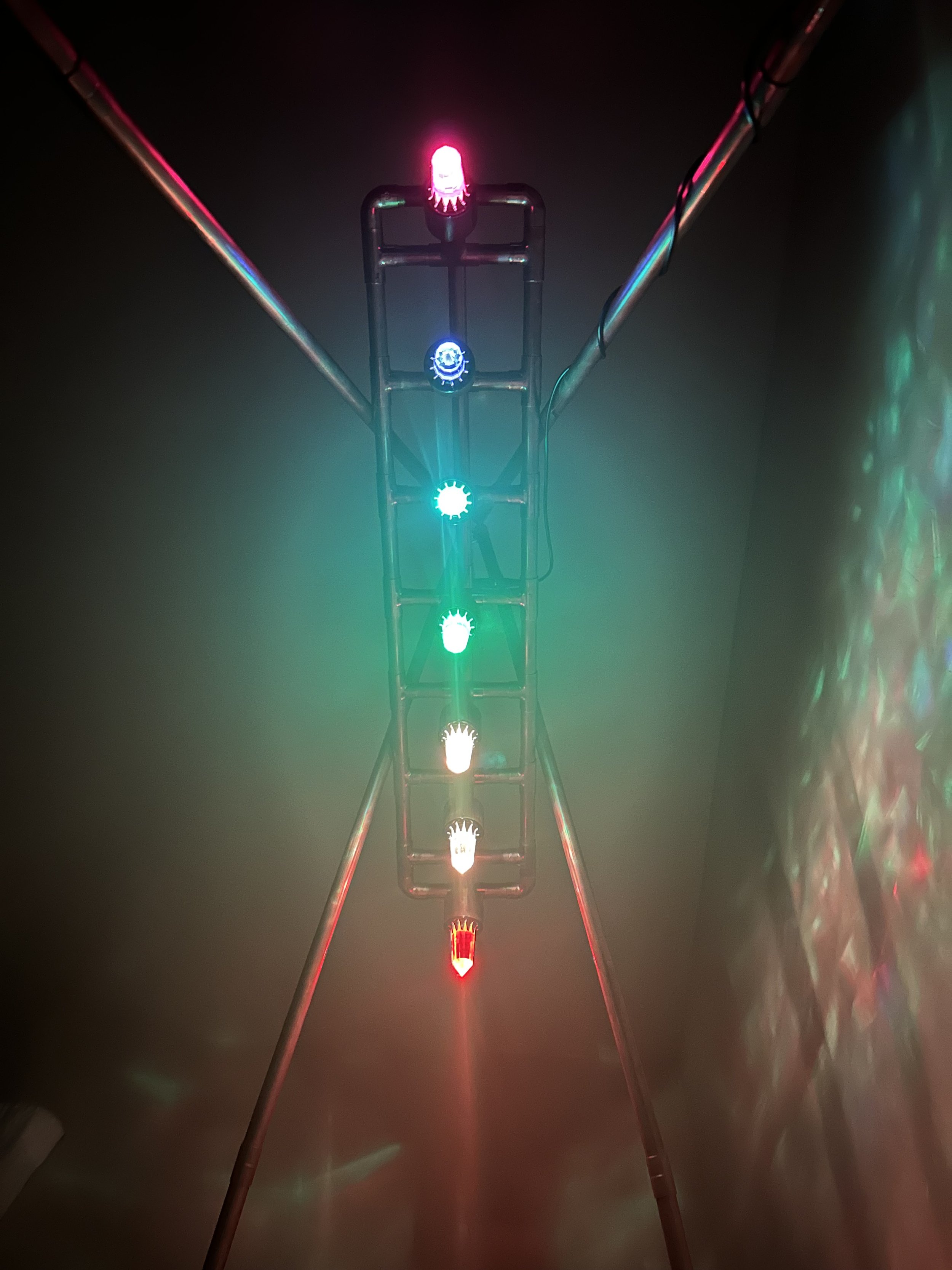 A tall, narrow structure with multiple multicolored  Vogel crystal lights, including Chakra-aligned red, orange, green, blue, and pink glowing in a dark room.