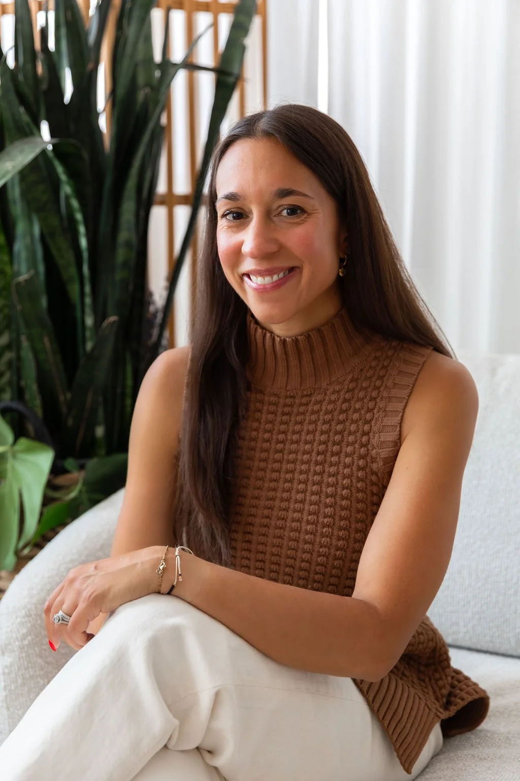 A woman with long brown hair, wearing a sleeveless brown sweater and white pants, sitting on a light-colored sofa with green plants in the background and sheer white curtains.