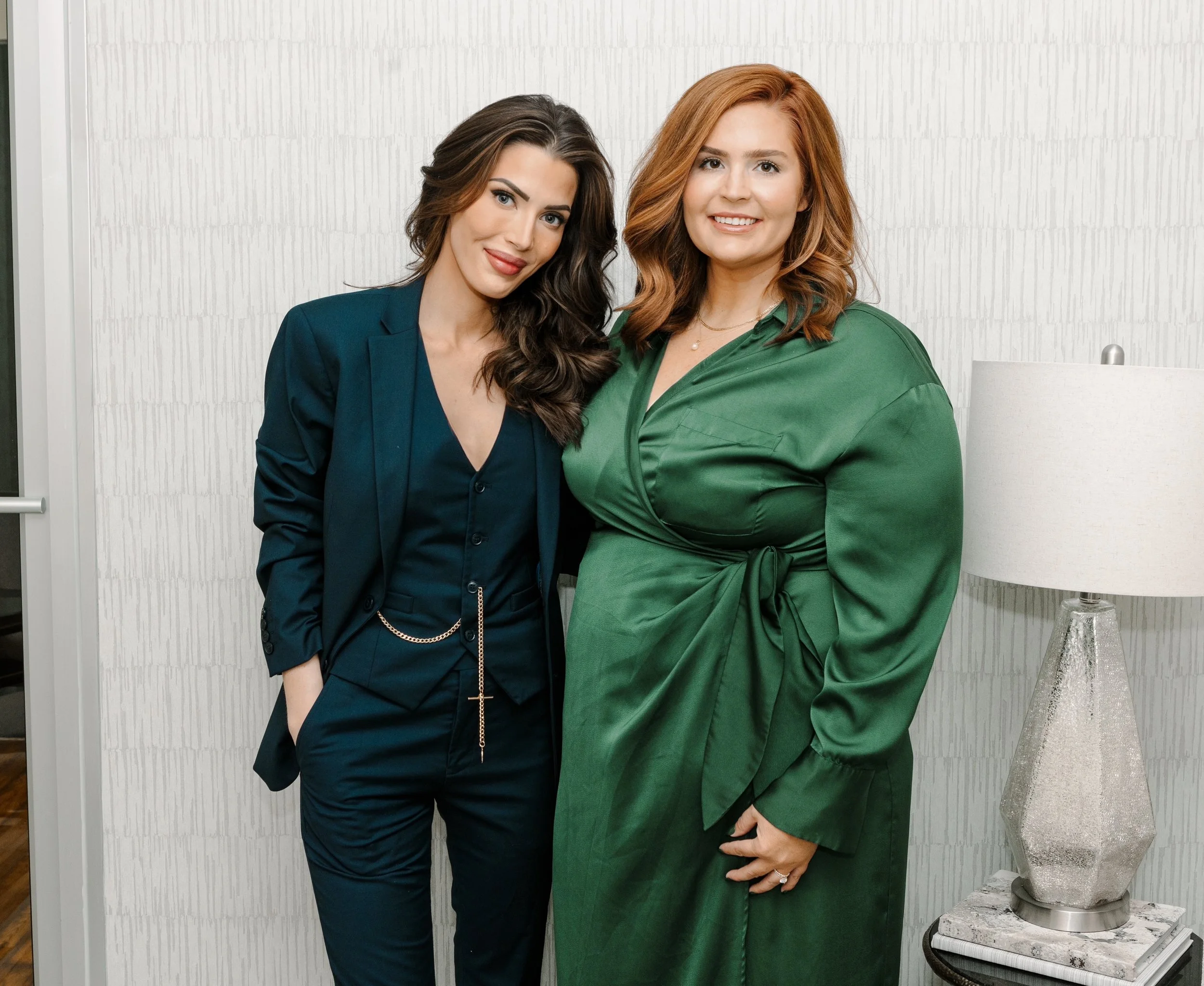 Two women standing together indoors, one in a dark green blazer and pants, and the other in a green satin wrap dress, smiling at the camera, with a white textured wall and a table lamp in the background.