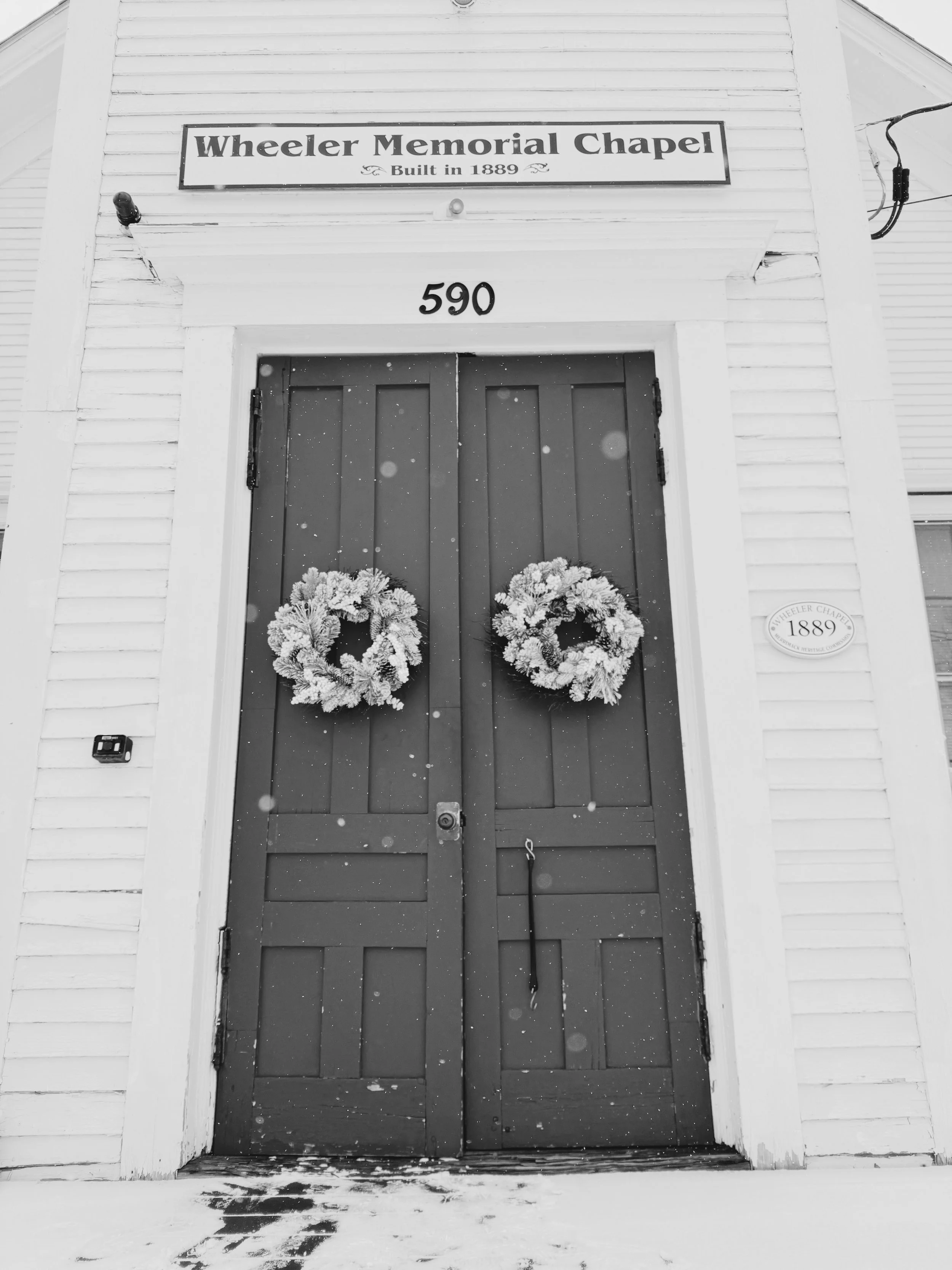 Black and white photo of the front entrance of Wheeler Memorial Chapel, built in 1889. Double wooden doors decorated with wreaths, with snow on the ground and falling snowflakes.