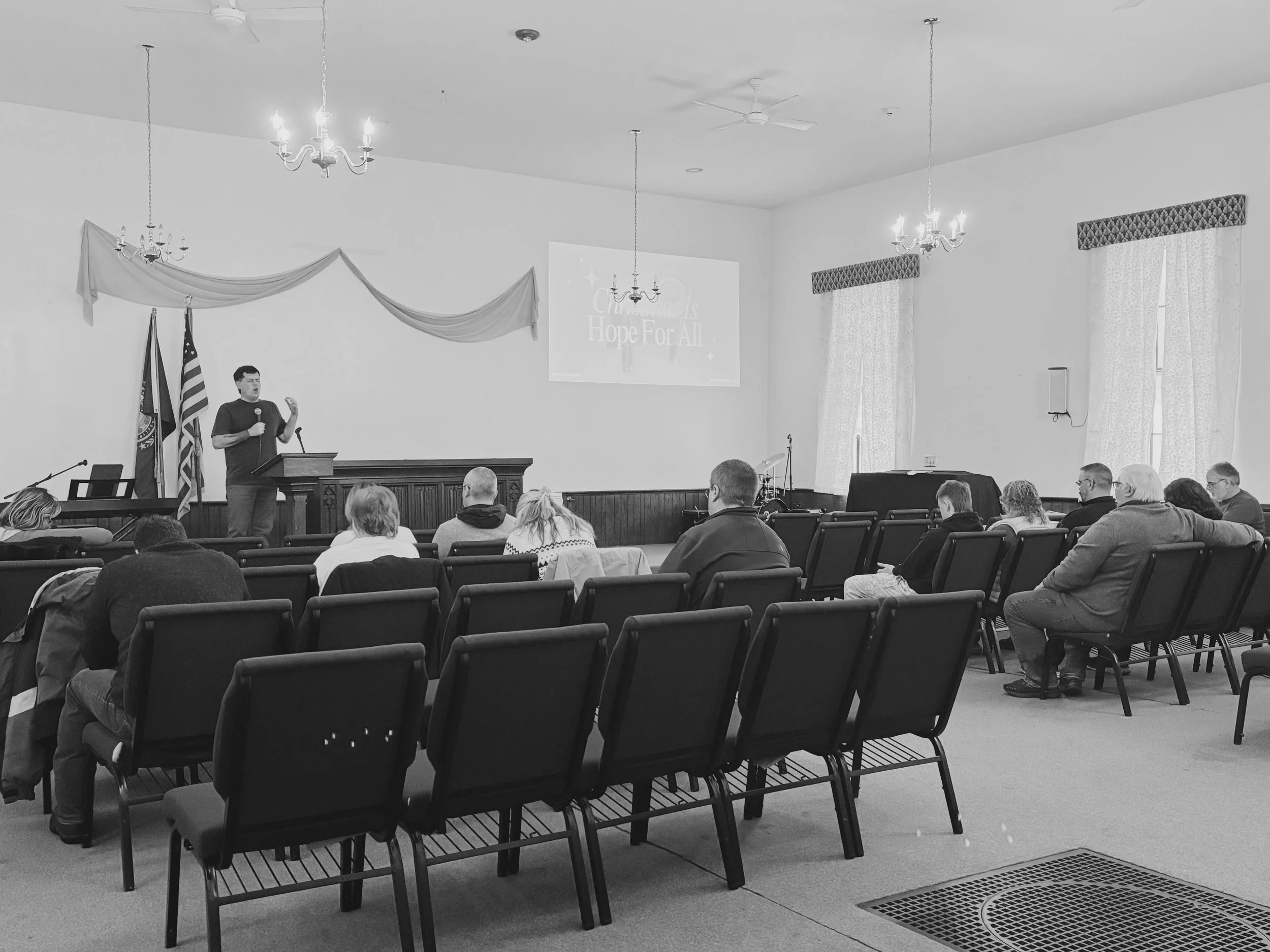 A man is giving a presentation or speech at a podium in a room with an audience seated in chairs, with a screen behind displaying the words 'Hope For All'. The room has curtains, chandeliers, and flags at the front.