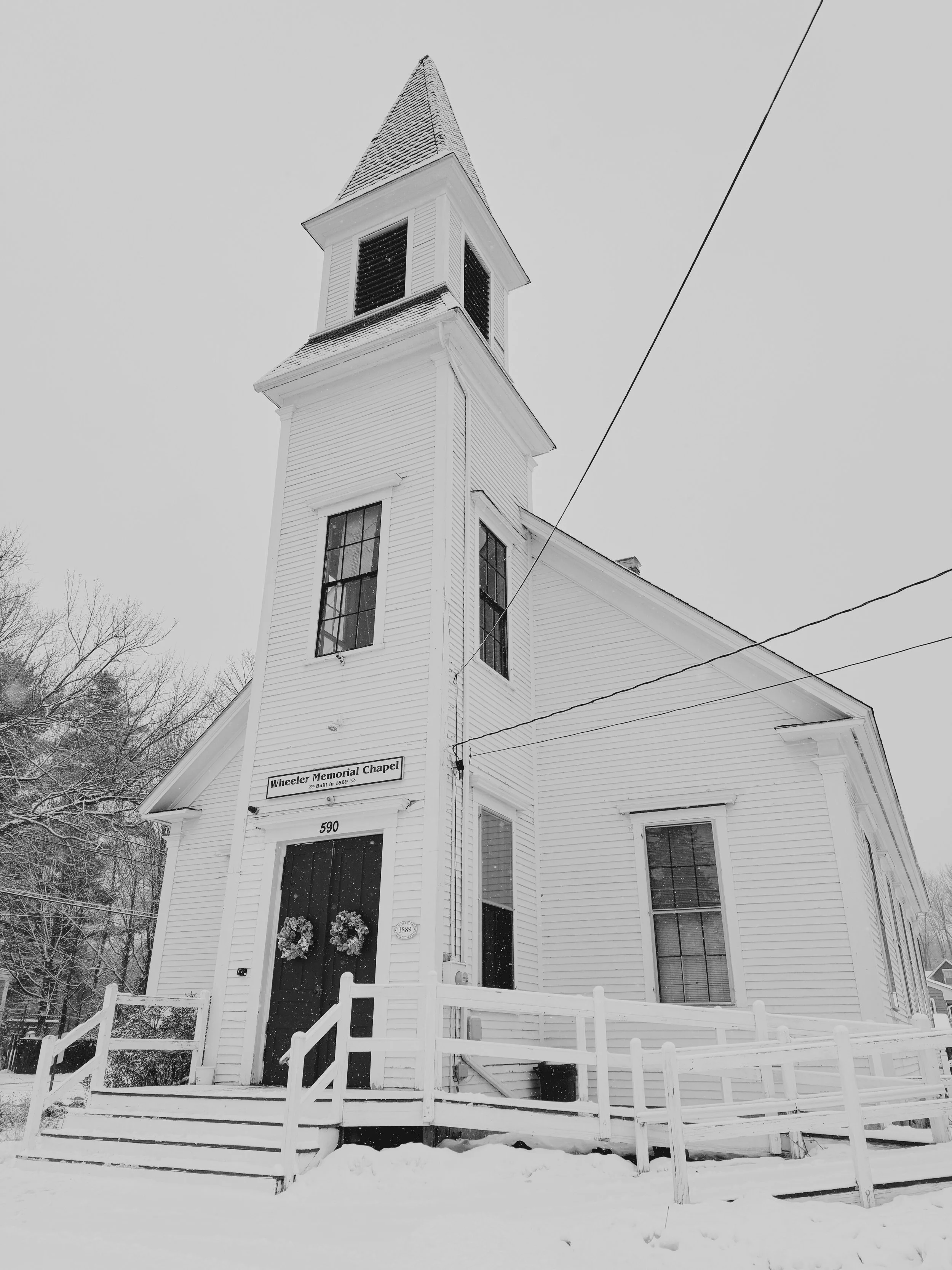 Black and white photo of Wheeler Memorial Chapel, a white wooden church with a tall steeple, snow on the ground and wreaths on the entrance door.