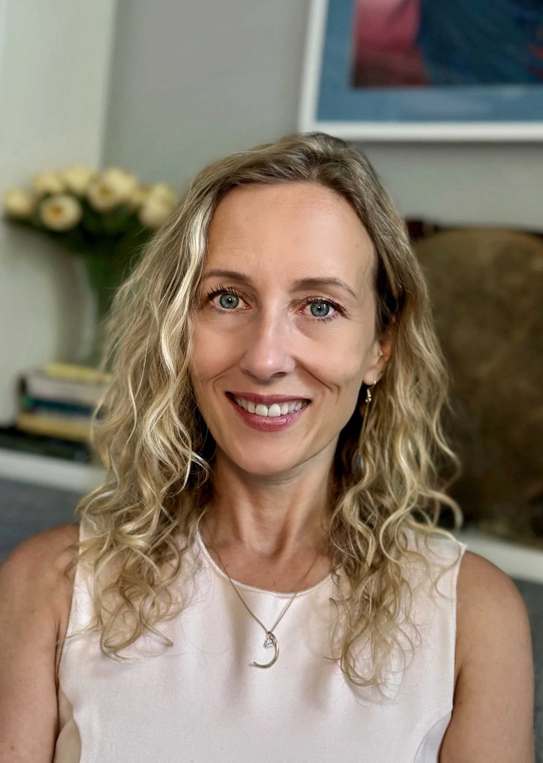A woman with curly blonde hair smiling, wearing a white sleeveless top, a necklace with a moon pendant, and earrings, sitting in a room with a vase of white flowers and an abstract painting in the background.