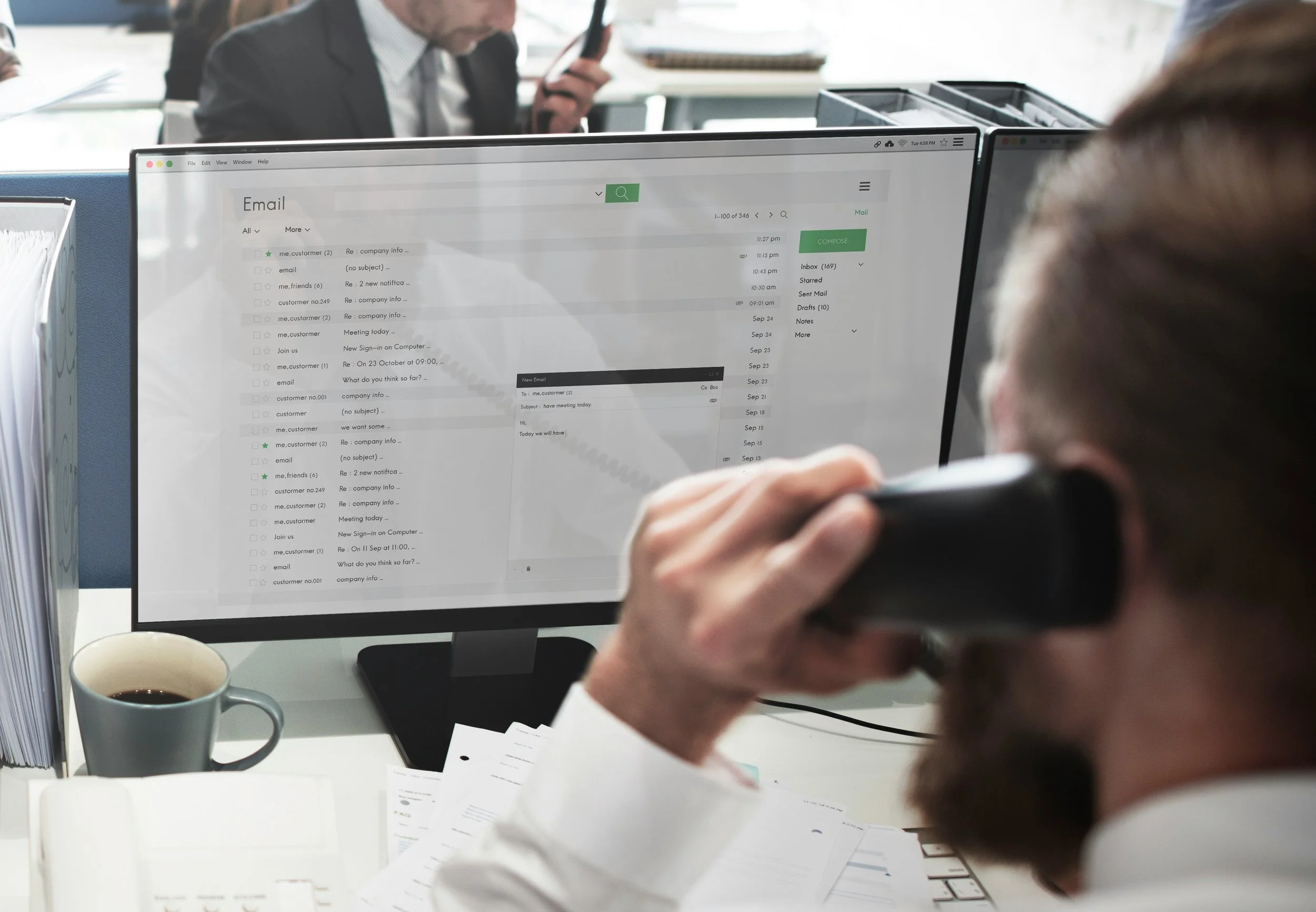 An office admin or dispatcher. He is on an office landline phone and staring at a monitor that shows an email list interface.