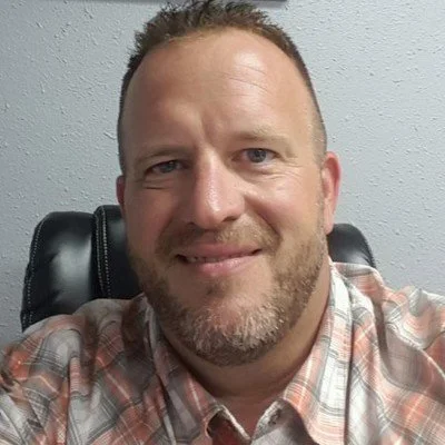 A smiling man with short hair and a beard, wearing a plaid shirt, sitting in front of a plain light-colored wall.