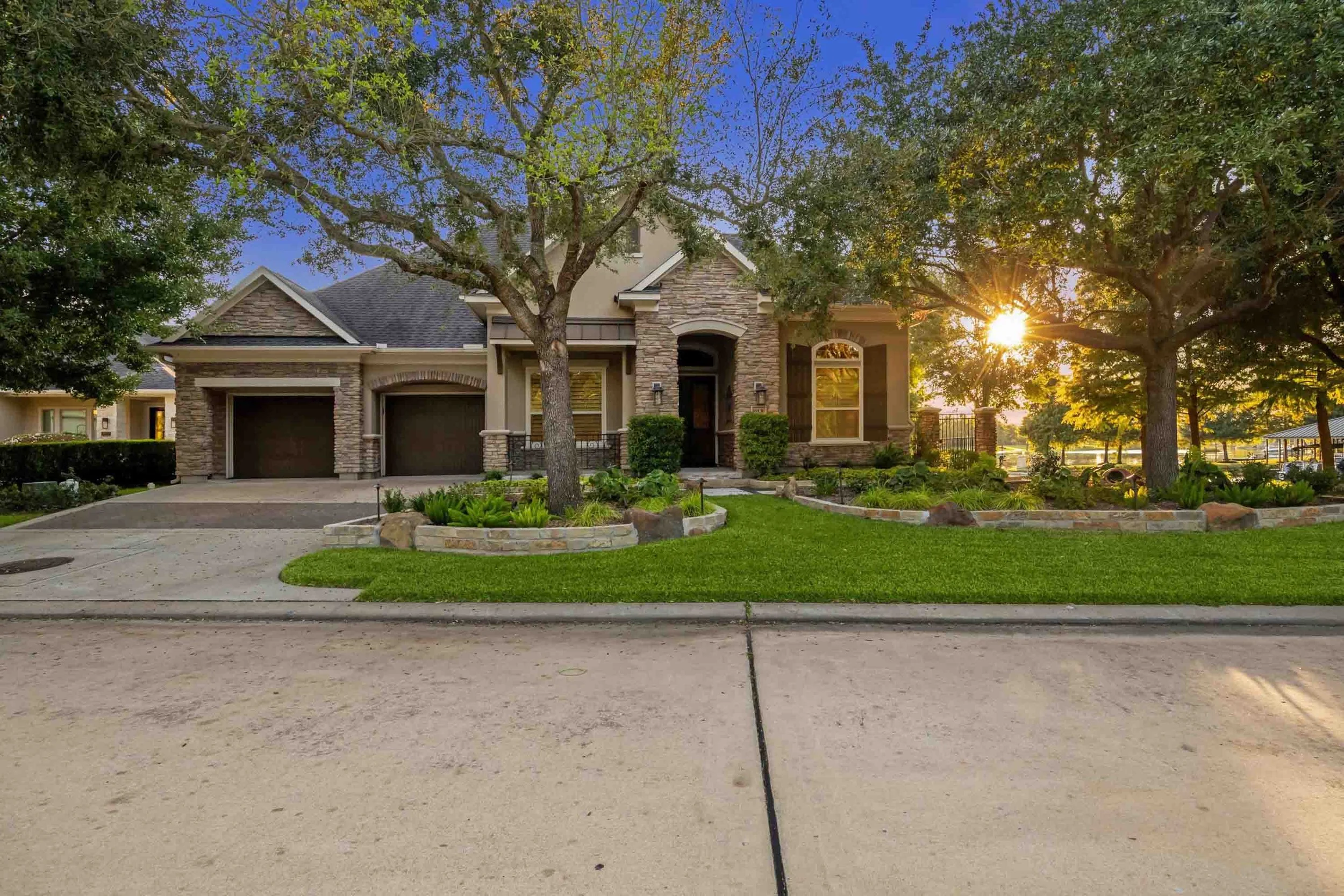 Brian Gardens Houston Landscaping - A modern house with a landscaped front yard, two trees, a driveway leading to a three-car garage, and the sun setting behind the trees.