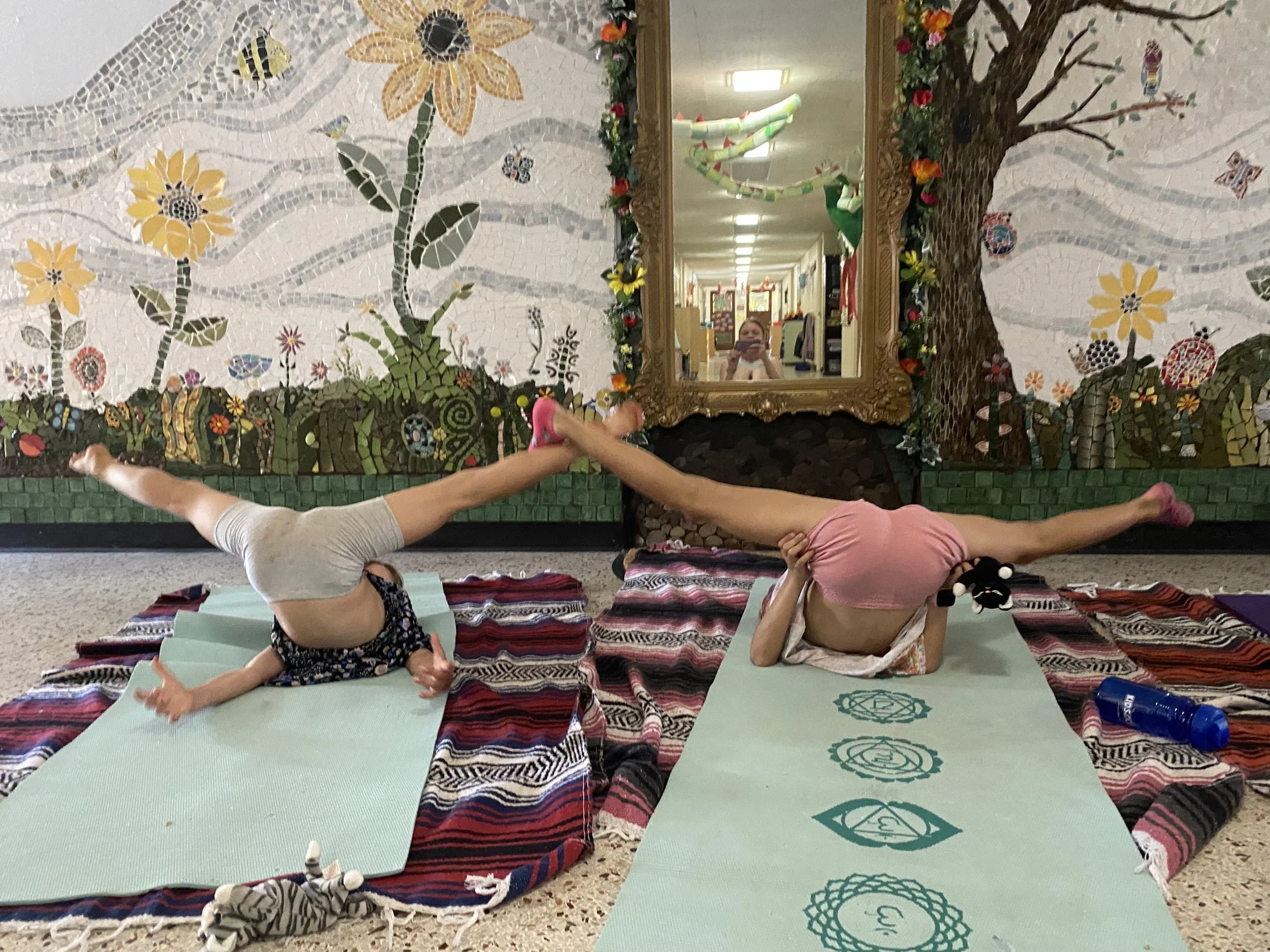 Two young girls performing yoga poses on mats in a decorated room with a colorful floral mosaic mural, a large ornate mirror, and a reflection of a person taking the photo.