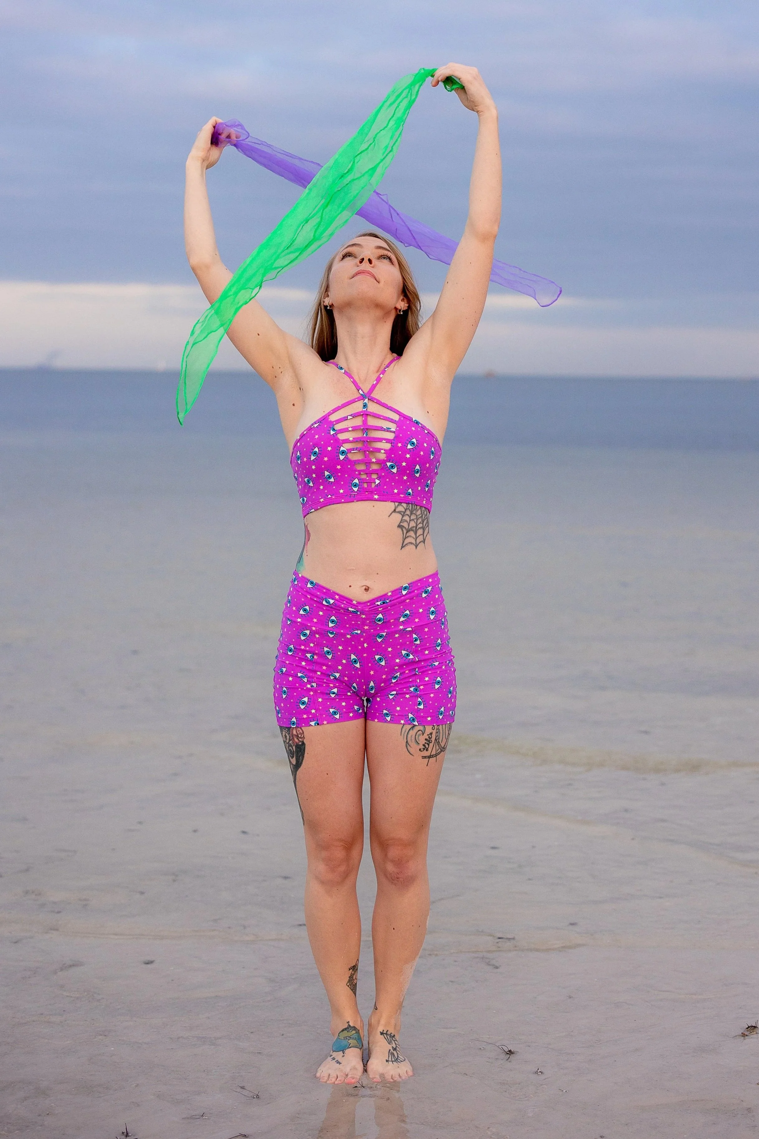 A woman standing on a beach with her arms raised, holding colorful sheer scarves, wearing a purple matching top and shorts with eye pattern, and having tattoos on her legs and torso, with the ocean and sky in the background.