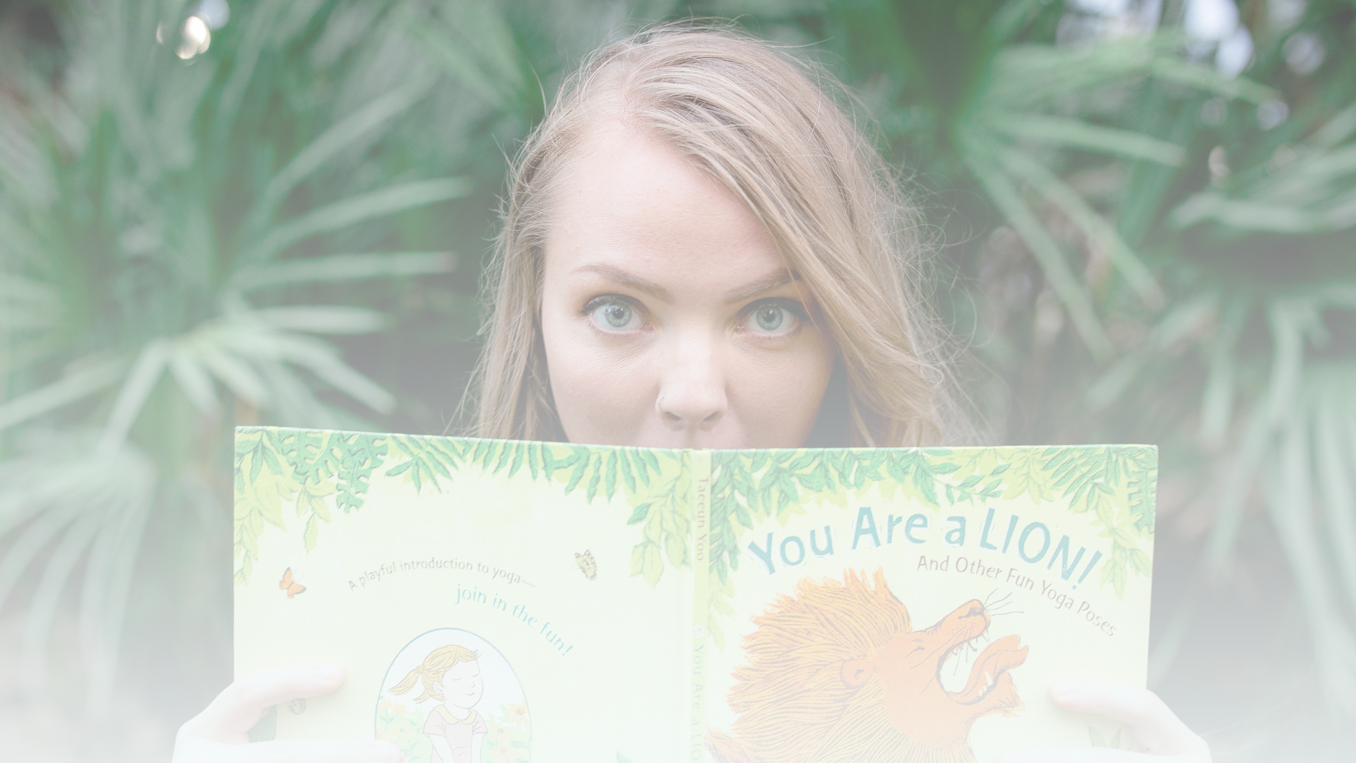 A woman with blue eyes and long, light brown hair peeks over a children's book titled "You Are a LION!" with a lion illustration on the cover, surrounded by green foliage in the background.