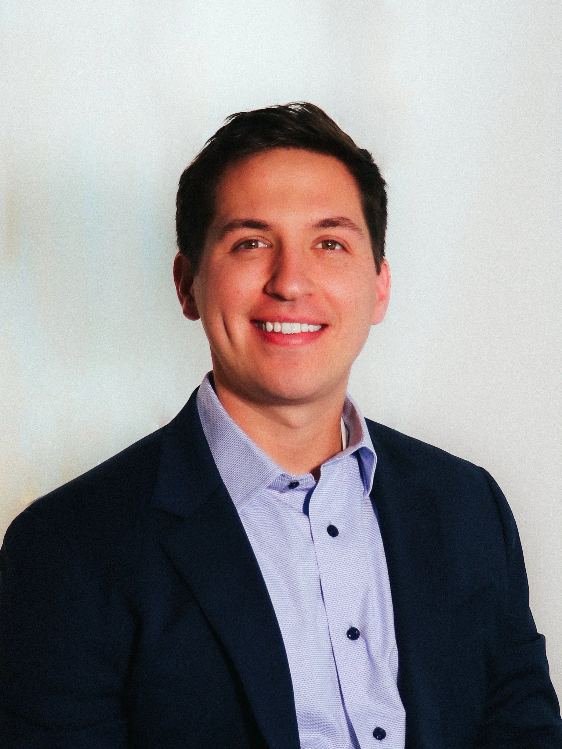 A young man with short dark hair, smiling, wearing a dark suit jacket and a light blue button-up shirt, standing against a plain white background.