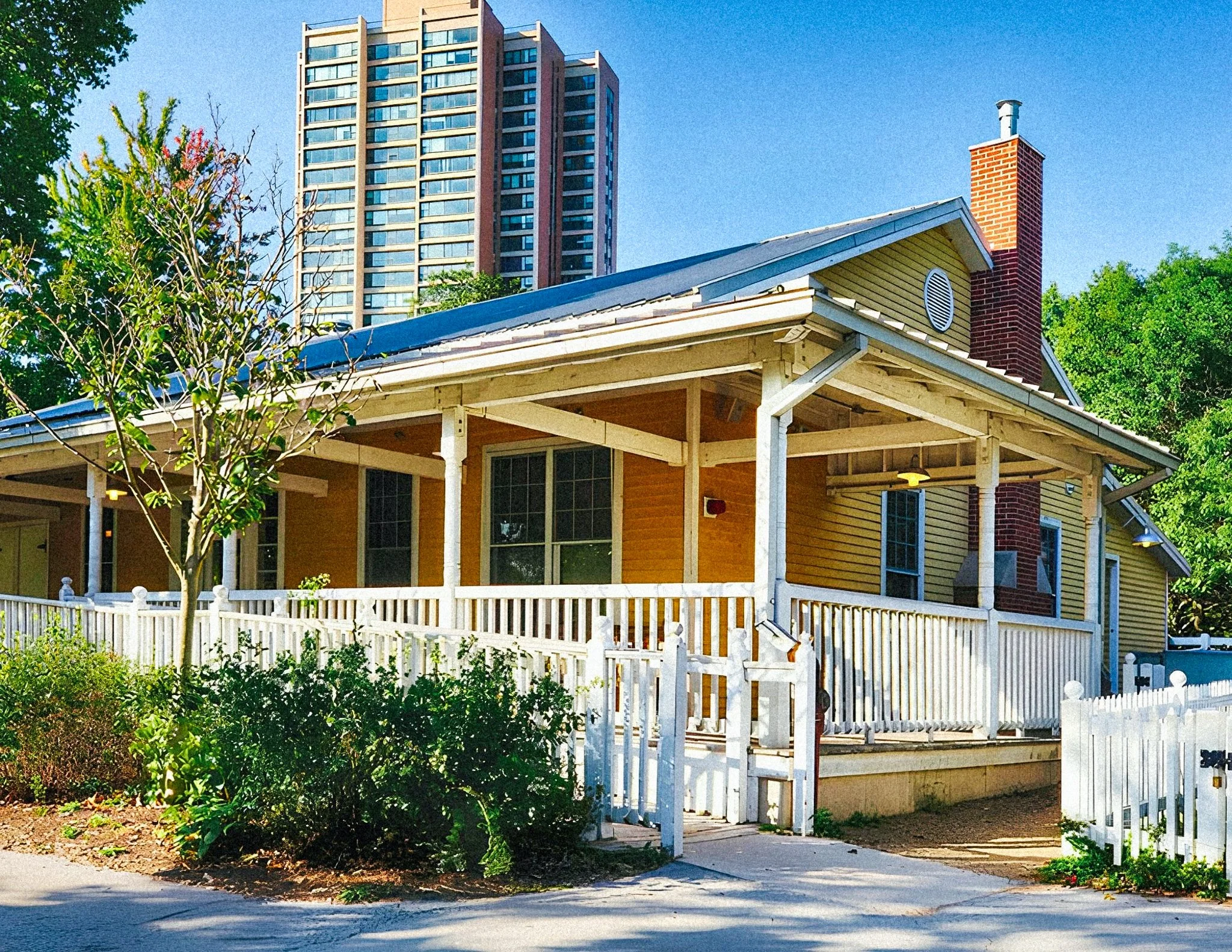 A house with a porch, white fence, yellow exterior, and red brick chimney, with trees and a high-rise building in the background against a blue sky.