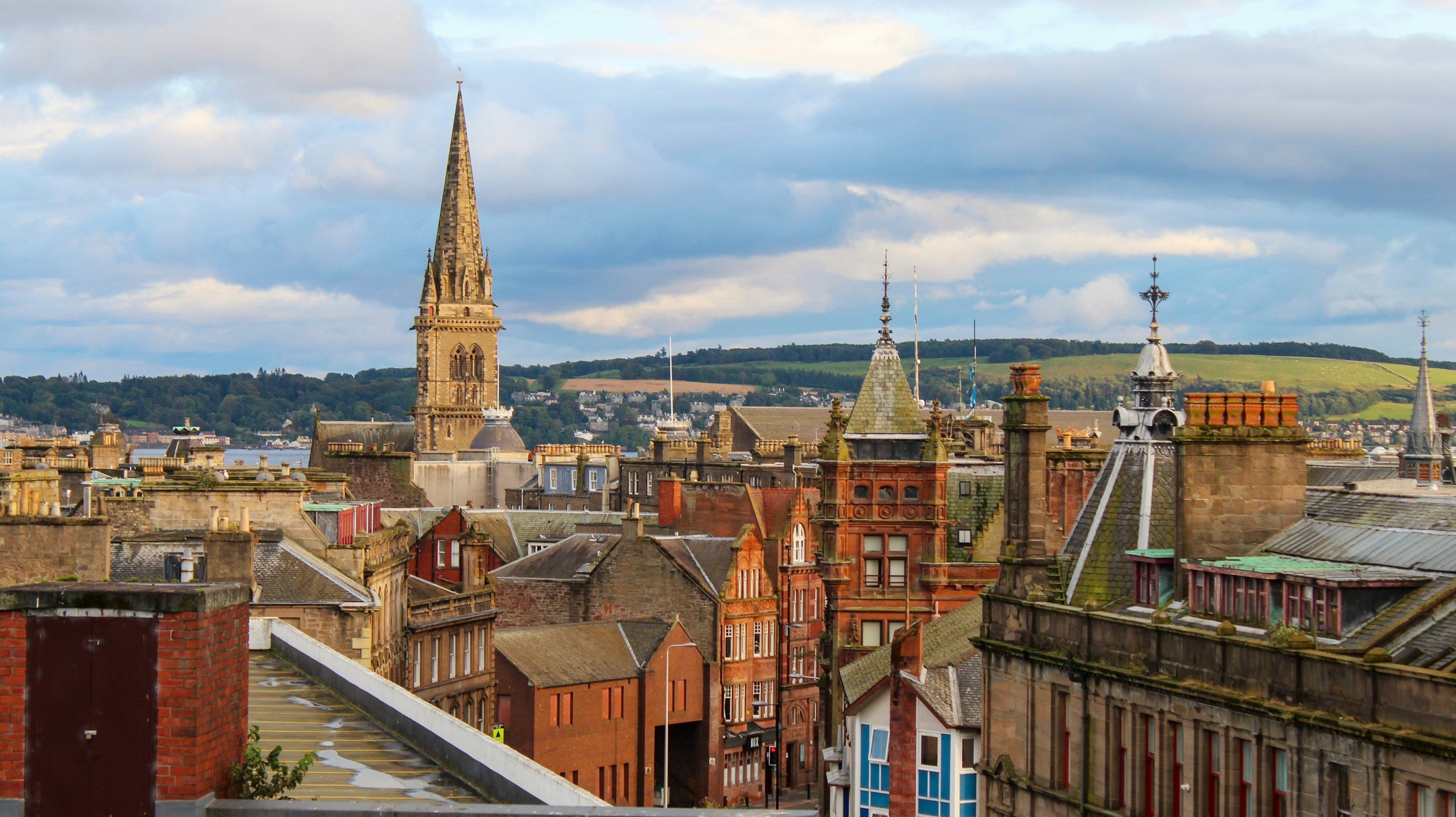 Cityscape with historic buildings and a prominent church spire, hills in the background, partly cloudy sky.