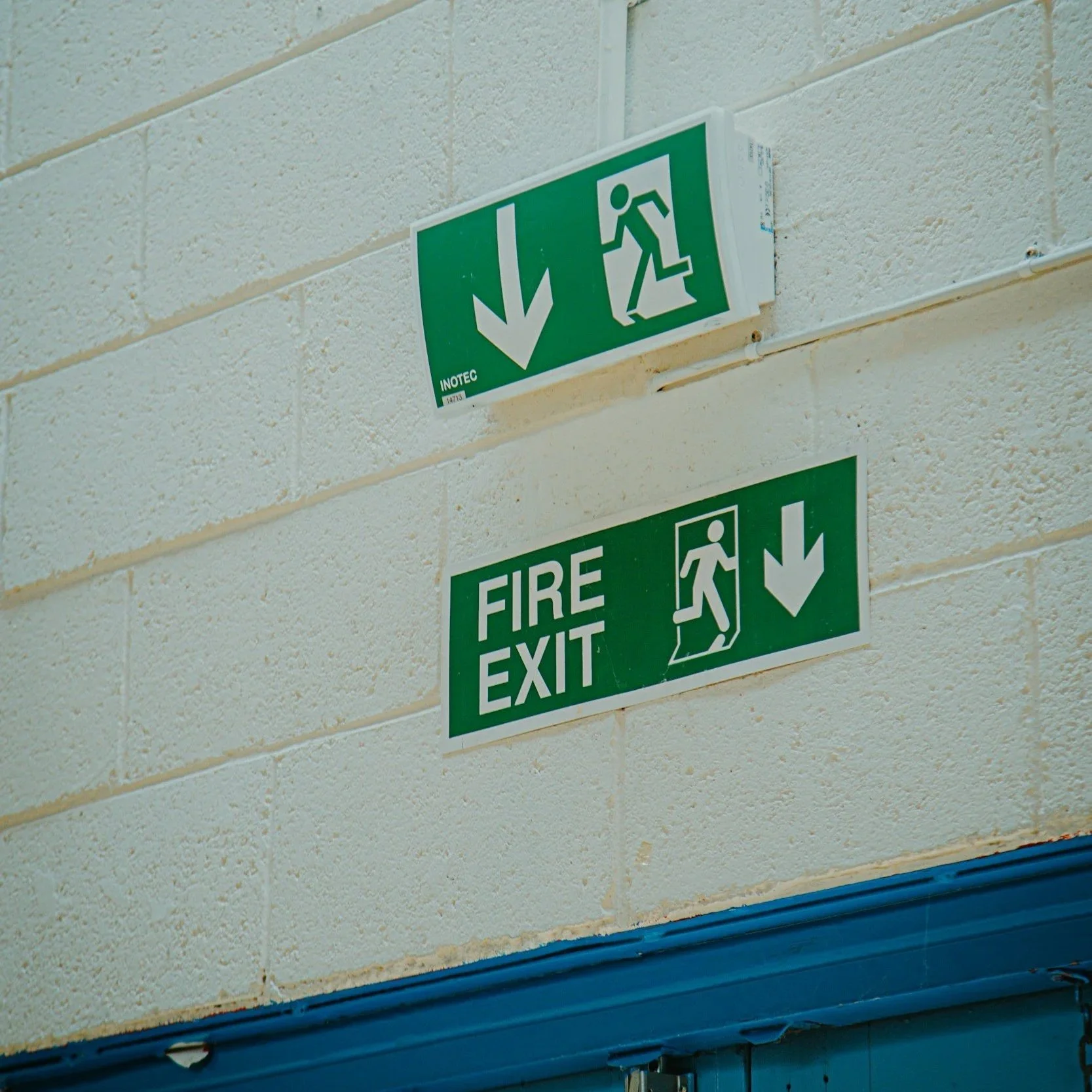 Green emergency exit signs on a beige brick wall, indicating directions for fire exit and emergency evacuation.