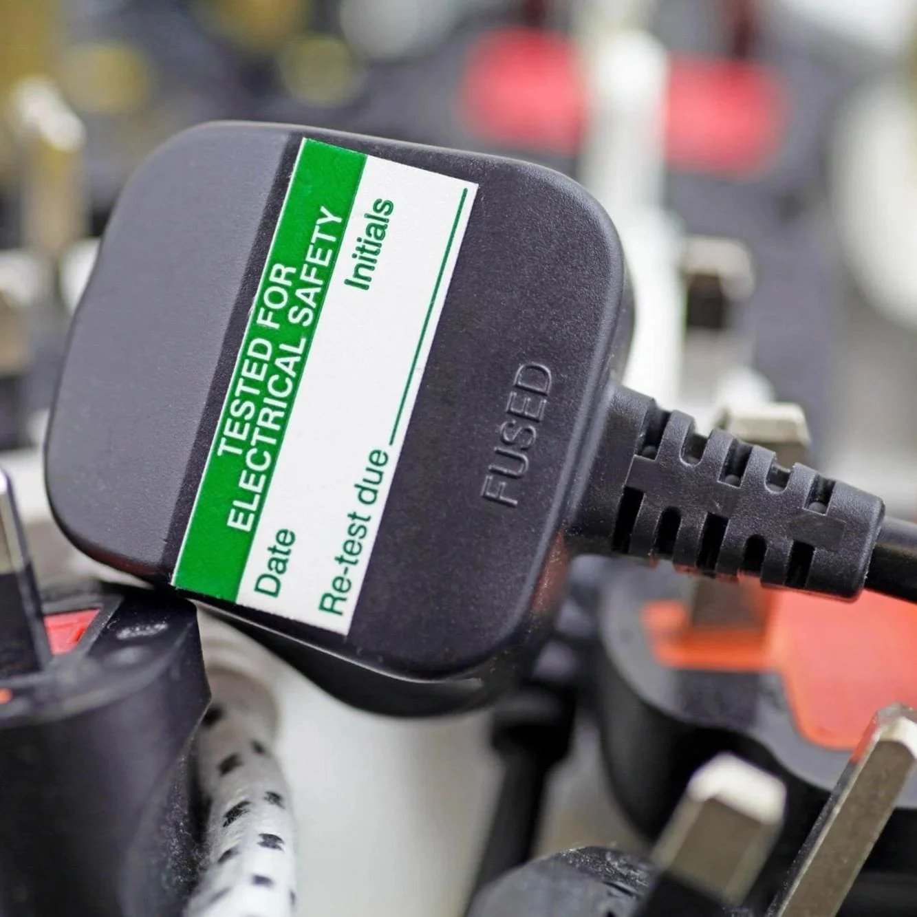 Close-up of an electrical safety inspection sticker on a power plug with the words 'Tested for Electrical Safety,' spaces for date and re-test due date, and blank initials, attached to protected outdoor electrical components.