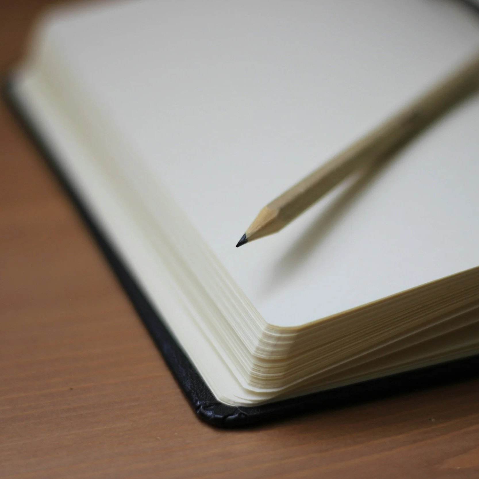 Open blank notebook with a sharpened wooden pencil resting on its pages, placed on a wooden surface.