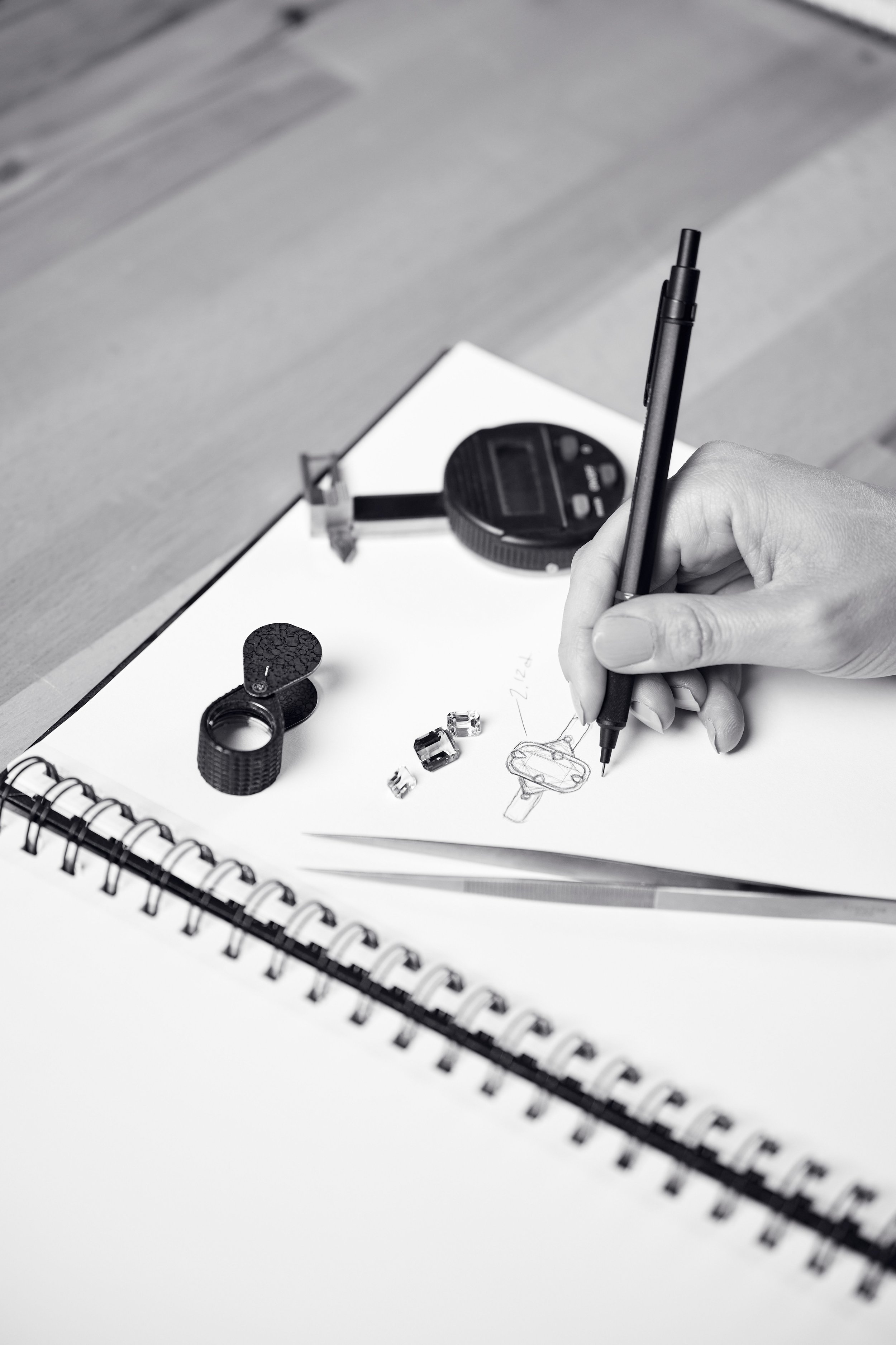 A person sketches a bike part on a notepad with a pen, with tools and small bike components on the desk, including a caliper, a gear, and a ring.