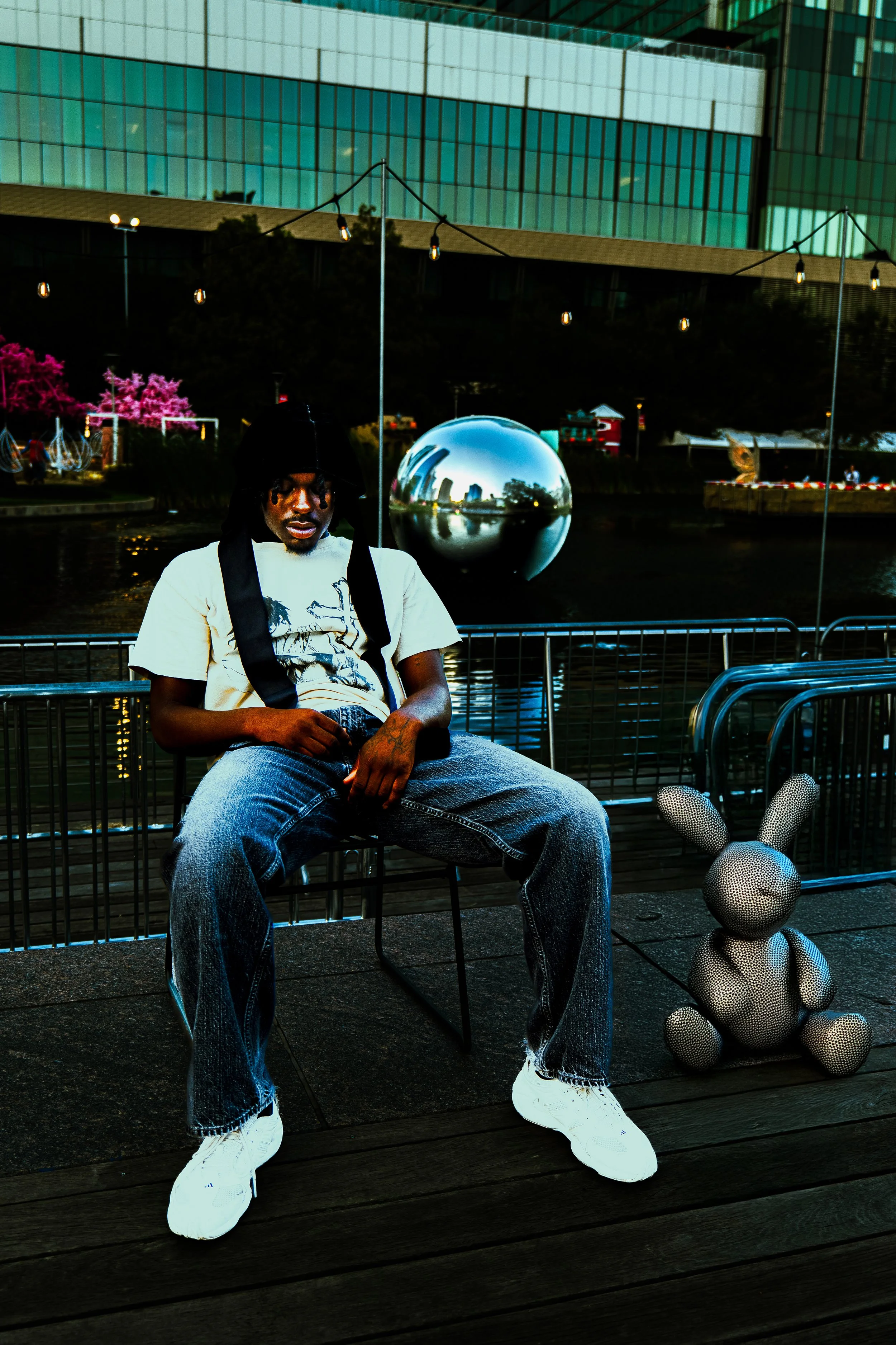 A man sitting on a chair outdoors near a body of water with a modern glass building in the background. There are string lights overhead, a sculpture of a bear, and a shiny metallic sphere reflecting the cityscape.