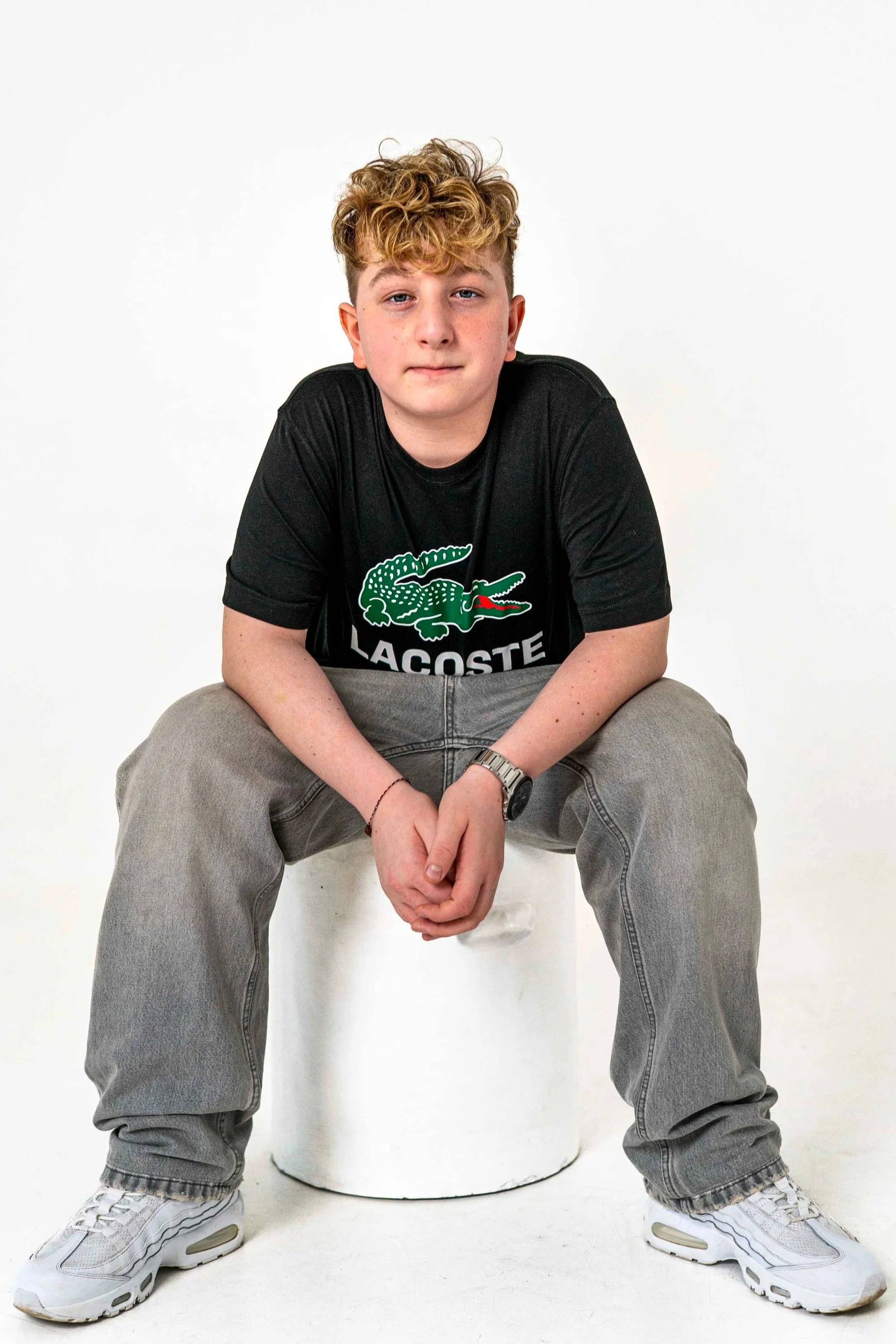 Young person with curly hair sitting on a white stool, wearing casual clothing including a black Lacoste T-shirt, gray pants, and white sneakers, against a plain white background.