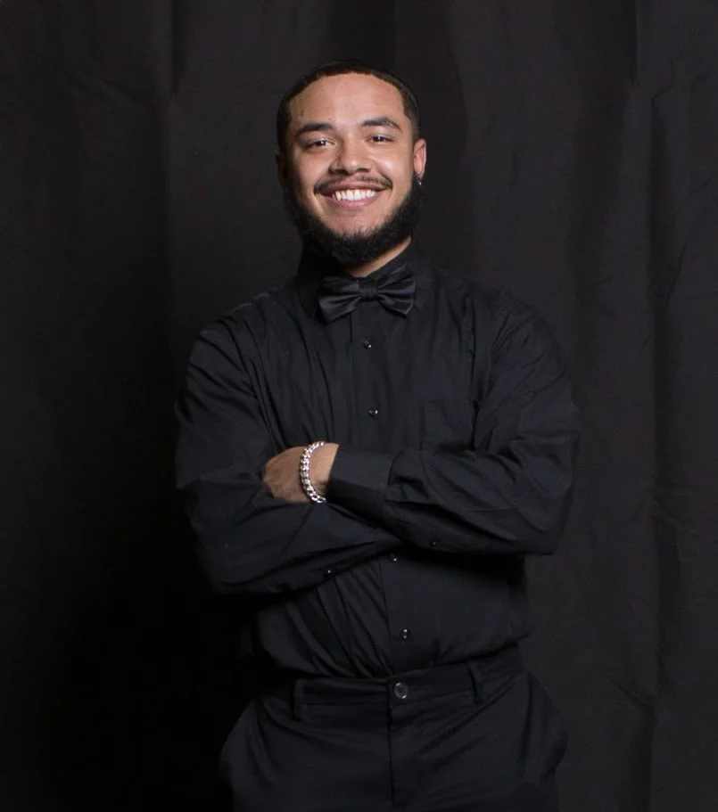 A smiling man with a beard, dressed in a black shirt and tuxedo with a bow tie, standing against a dark background with arms crossed.
