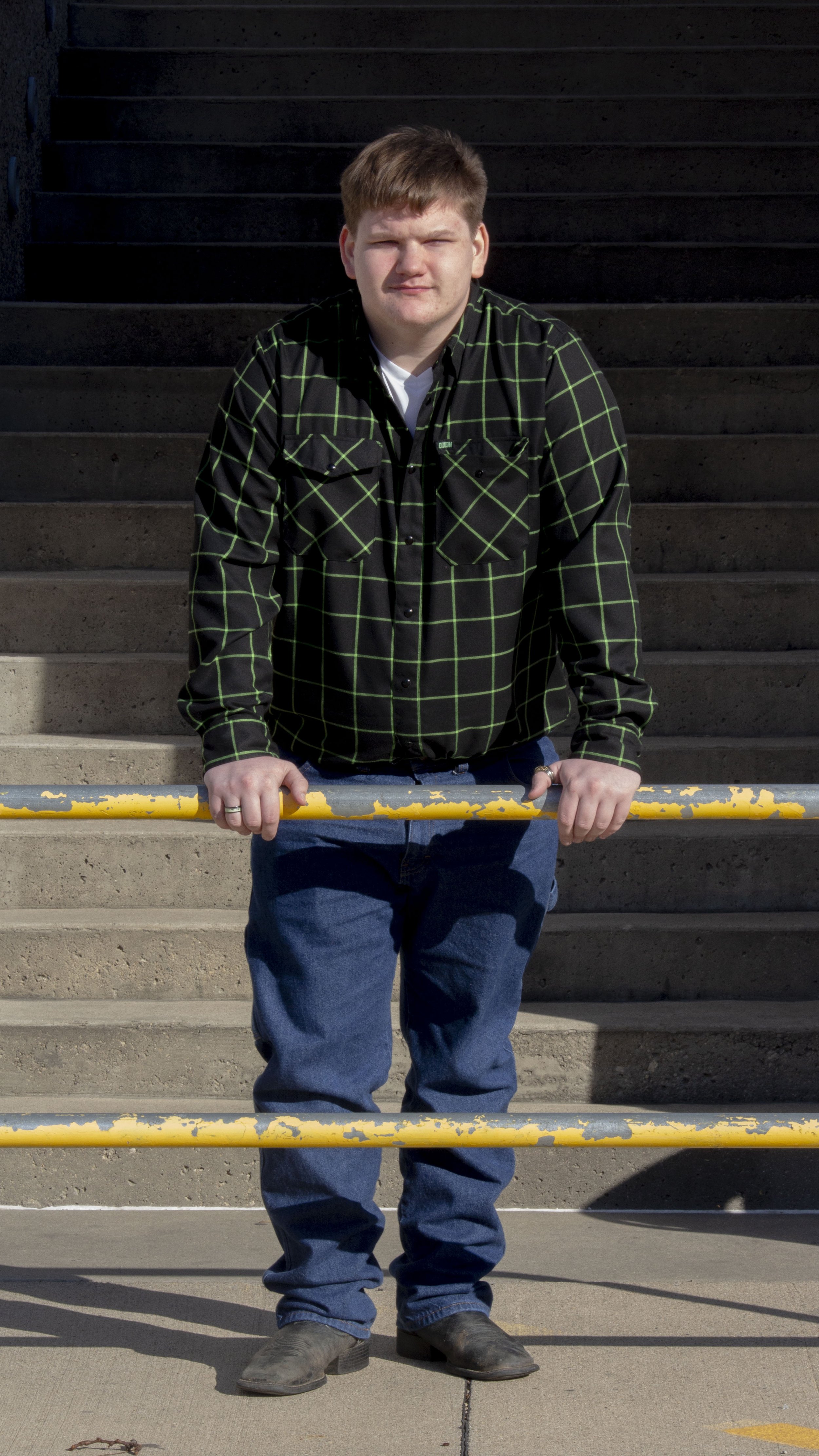 A young man with brown hair wearing a black and green checkered shirt, blue jeans, and gray boots, standing on a sidewalk in front of concrete steps, holding a yellow and gray metal bar.