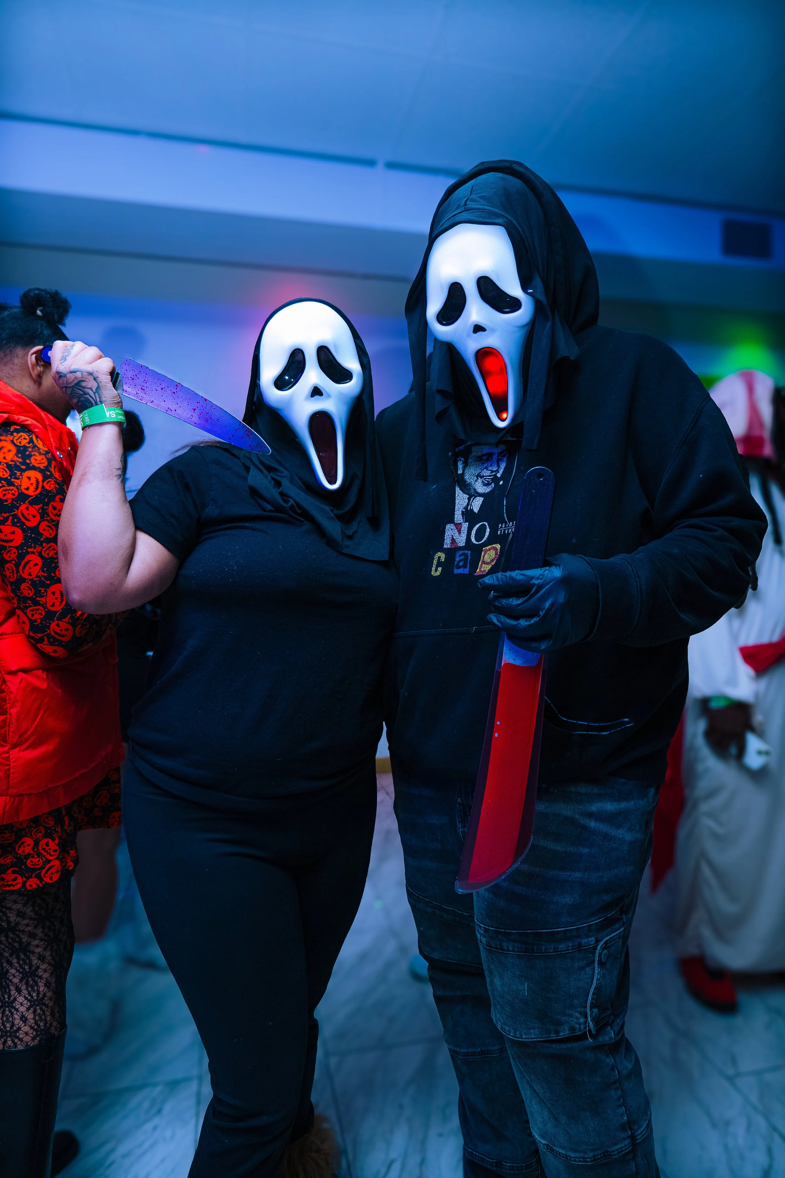 Two people wearing white ghost masks with black eye and mouth holes, dressed in dark clothing, stand close together with one holding a large prop knife with a red blade. The background is a dimly lit room with colorful lights and other people in cost