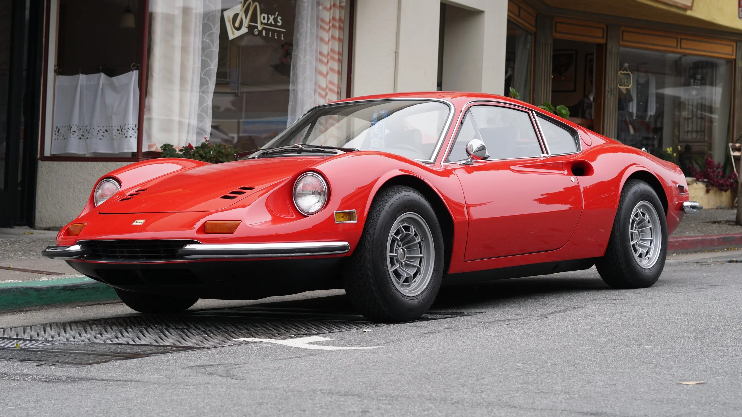 Red vintage sports car parked on city street in front of a restaurant with white curtains and potted plants outside.