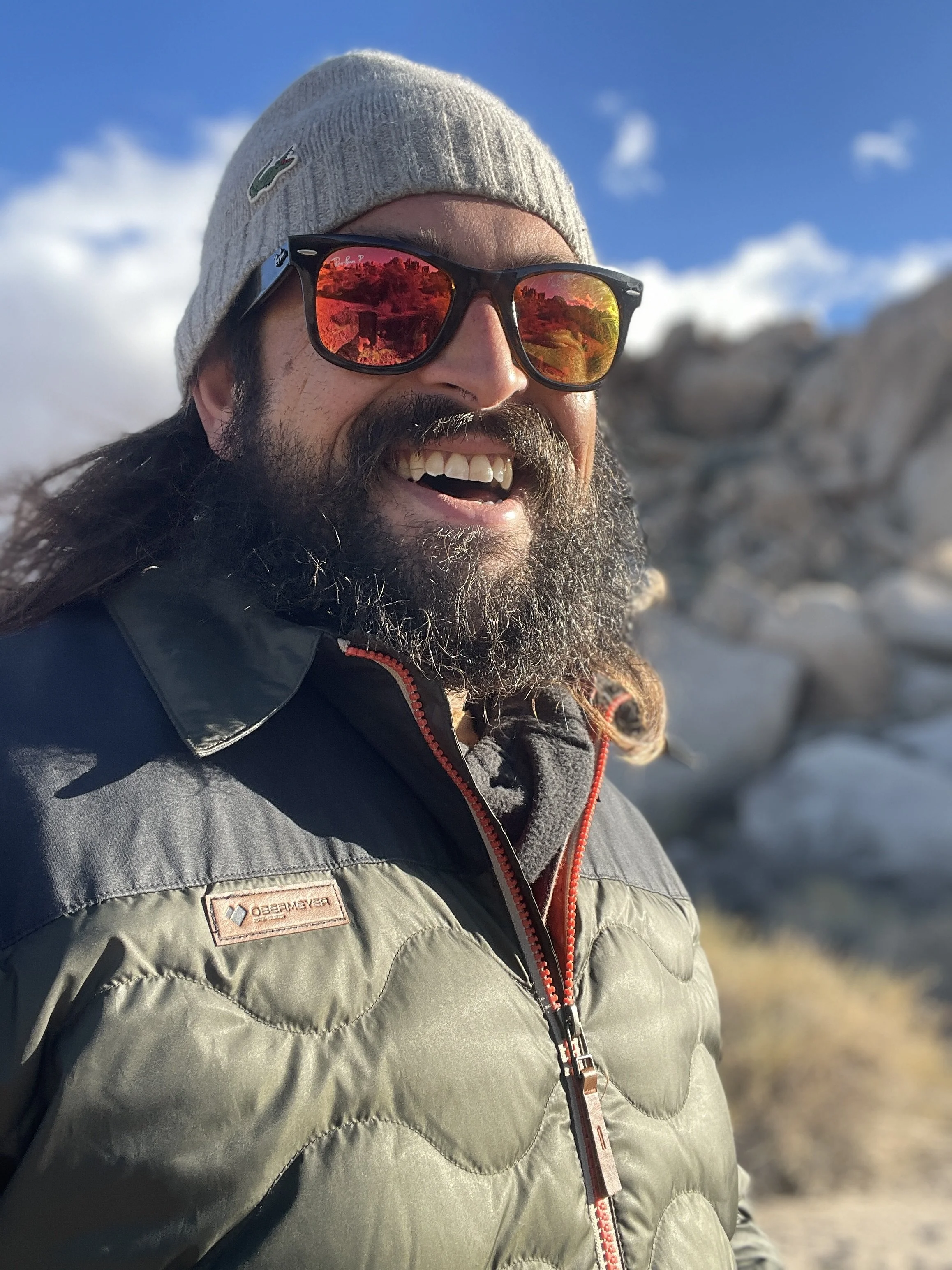 A man outdoors wearing a gray beanie, sunglasses with reflective red lenses, a gray and black jacket, smiling at the camera with snow and rocks in the background.