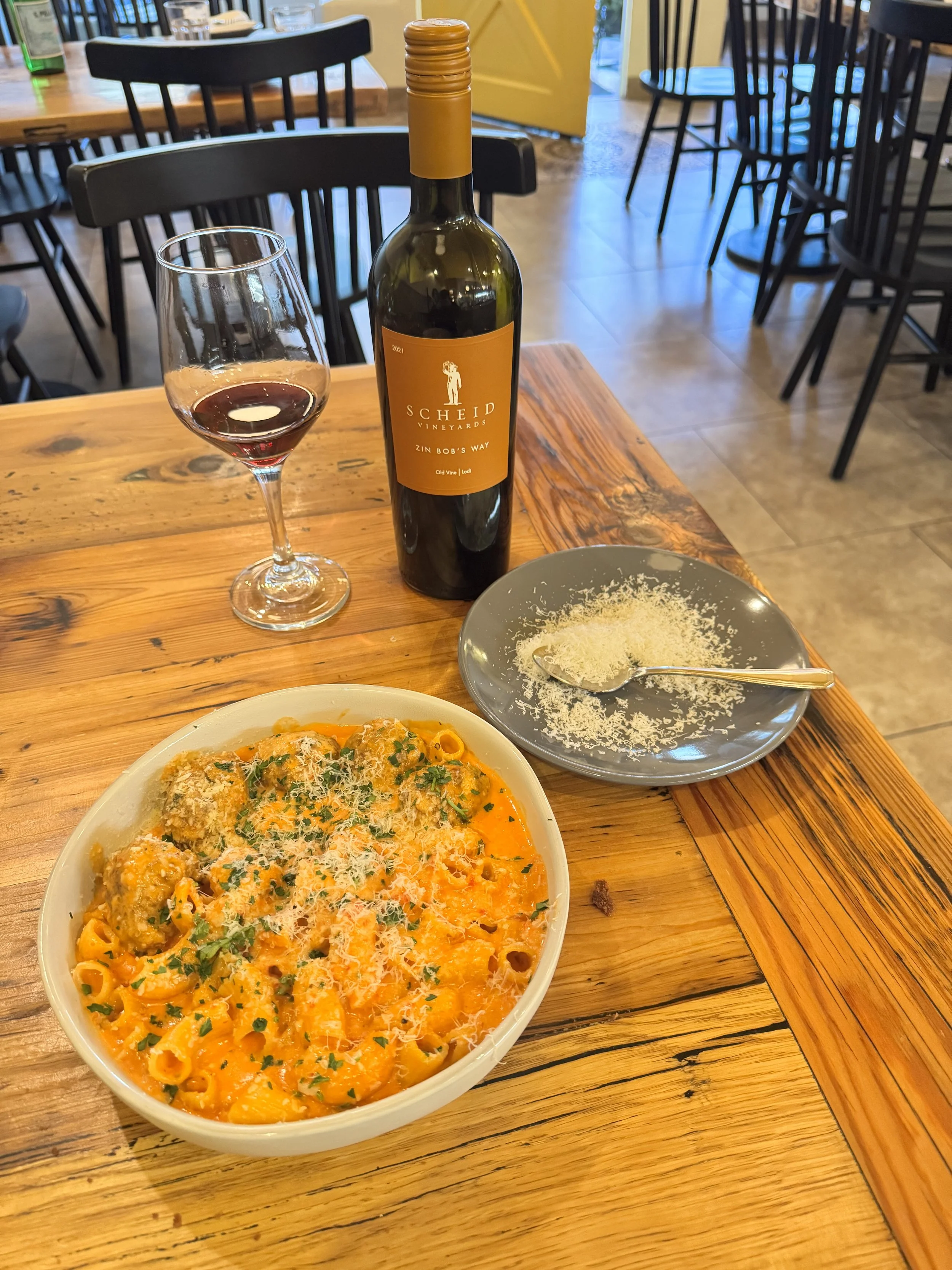 A glass of red wine, a bottle of Scheid Vineyards wine, and a baked pasta dish topped with cheese and parsley on a rustic wooden table in a restaurant.
