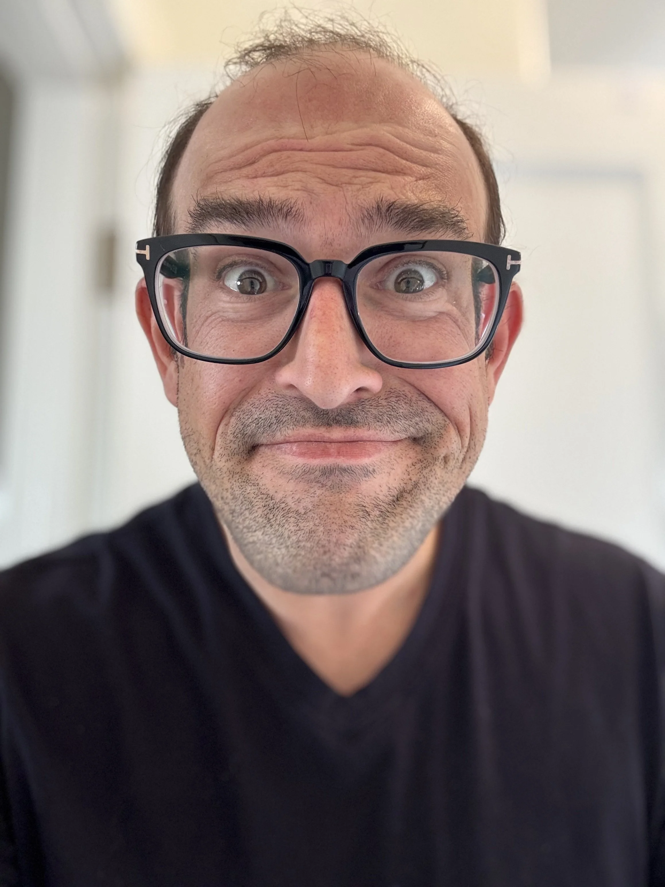 A man with glasses making a humorous facial expression, smiling with wide eyes and raised eyebrows.