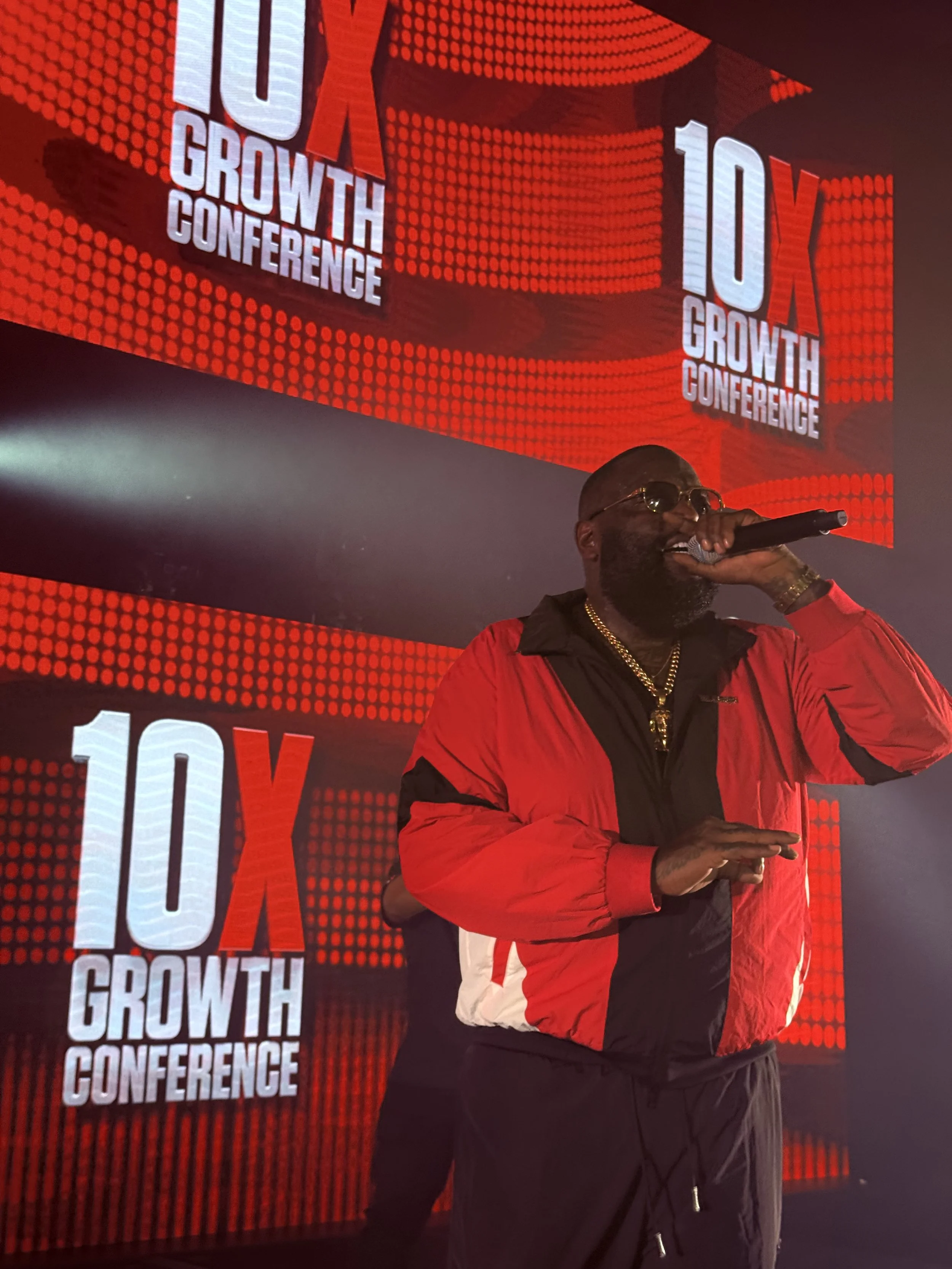 A man wearing sunglasses and a red and black jacket speaking into a microphone on stage at the 10X Growth Conference, with large digital screens displaying the event name behind him.