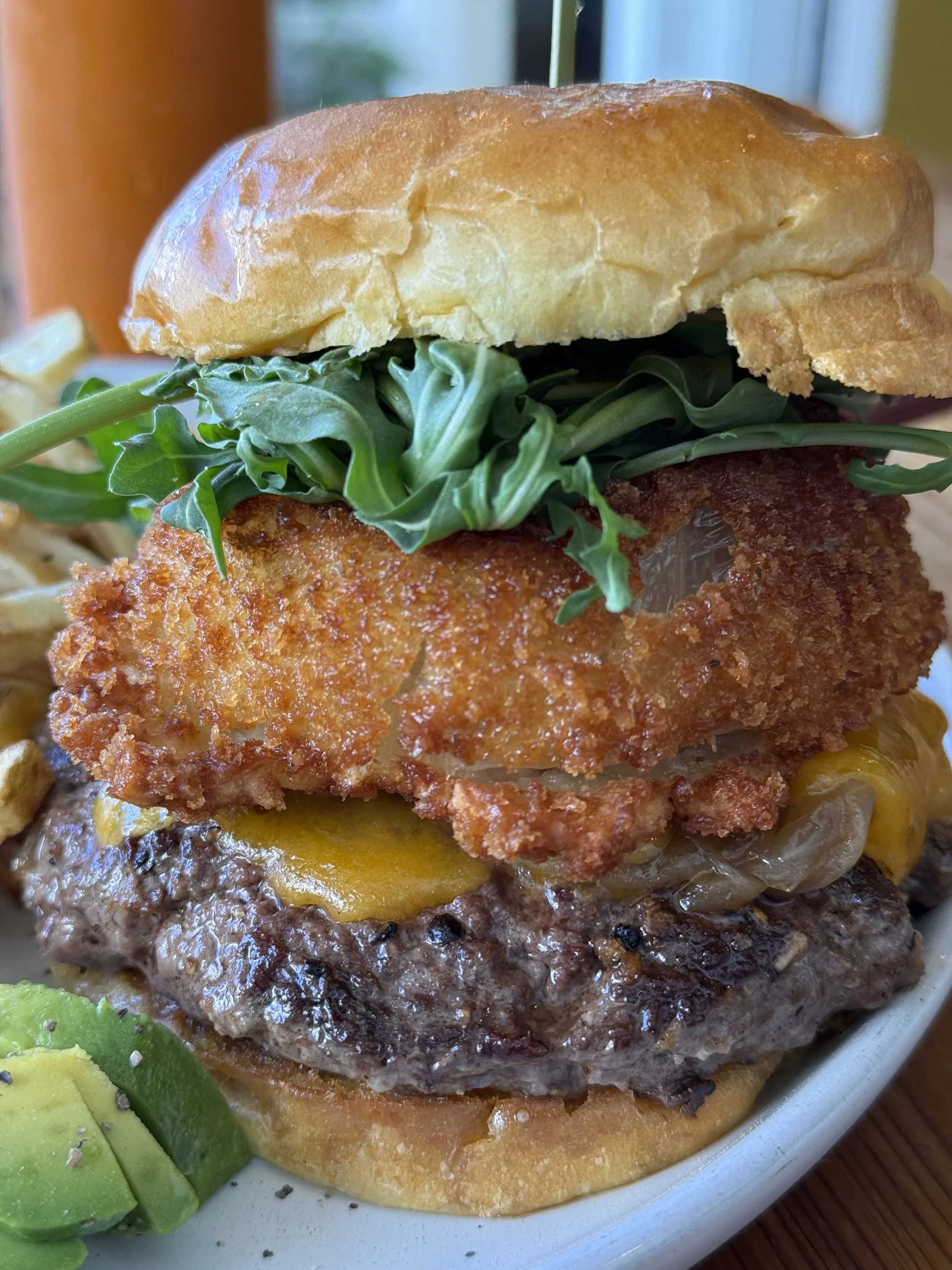 Close-up of a burger with a fried fish fillet, cheddar cheese, lettuce, and a toasted bun, served with sliced avocado on the side.
