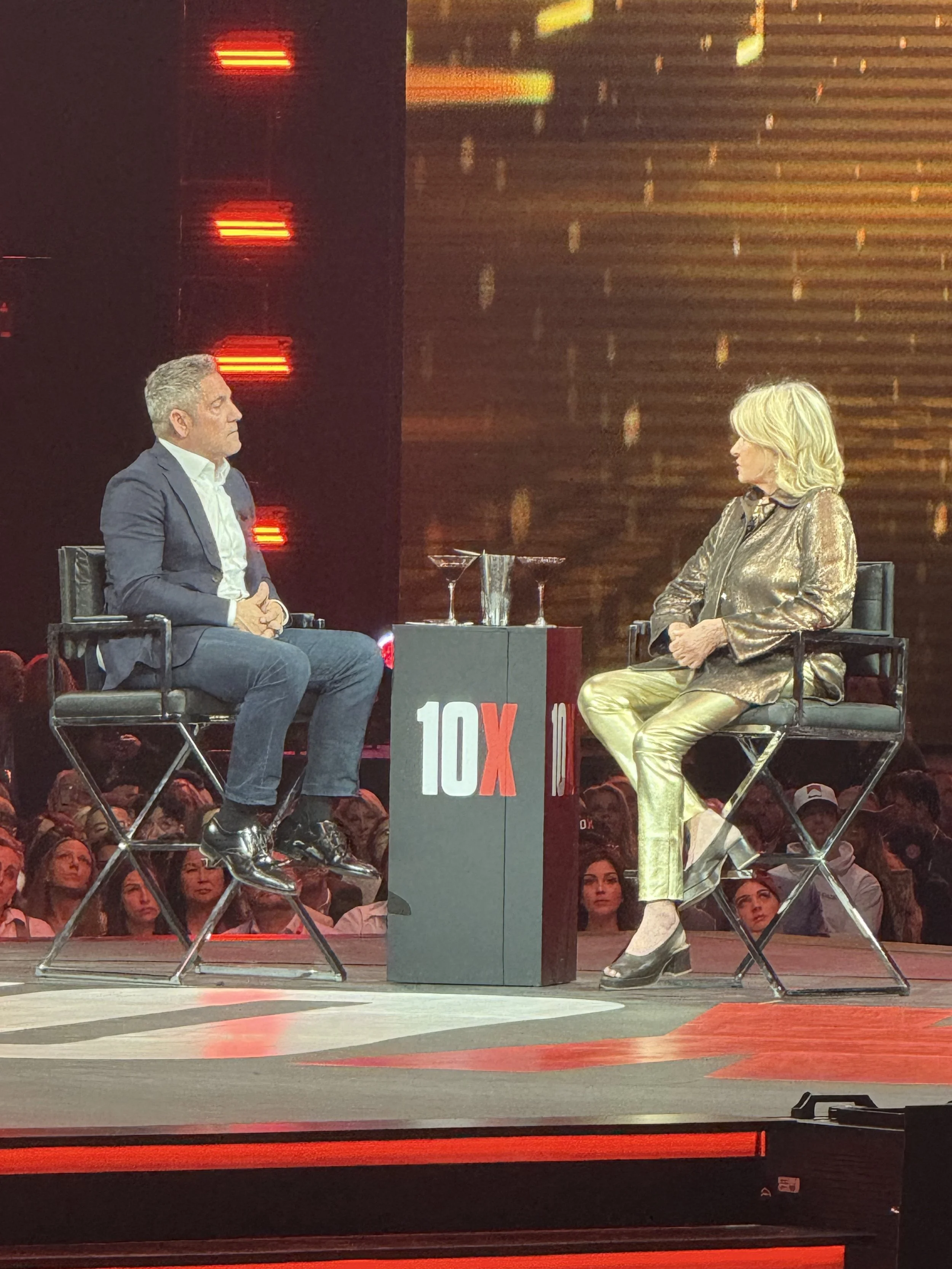 Two people, a man and a woman, sit on stage during a televised interview, with a '10X' logo on the table between them, and an audience visible in the background.