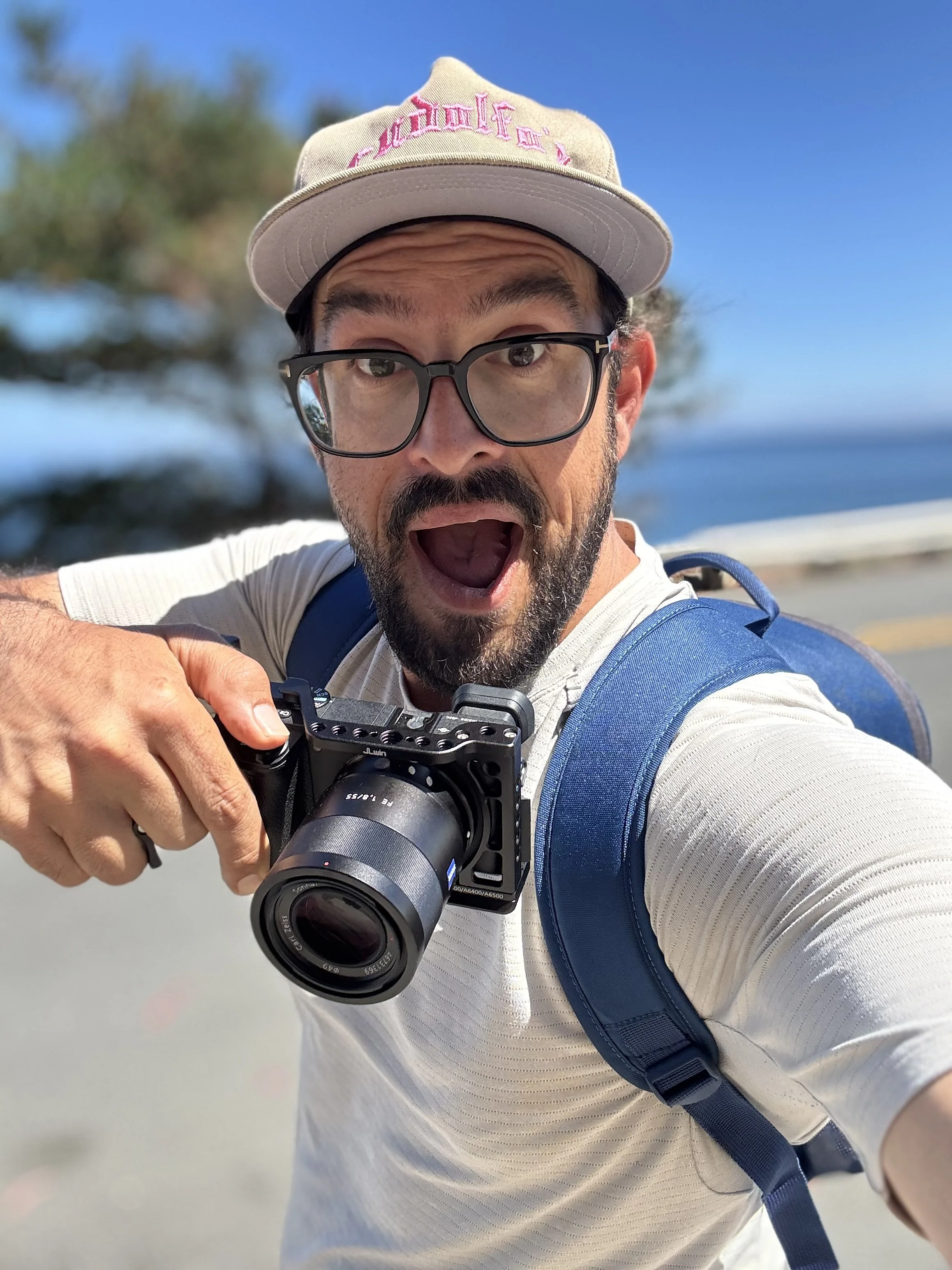 A man with glasses, a beard, and a baseball cap taking a selfie with a camera outdoors on a sunny day, with trees and the ocean in the background.