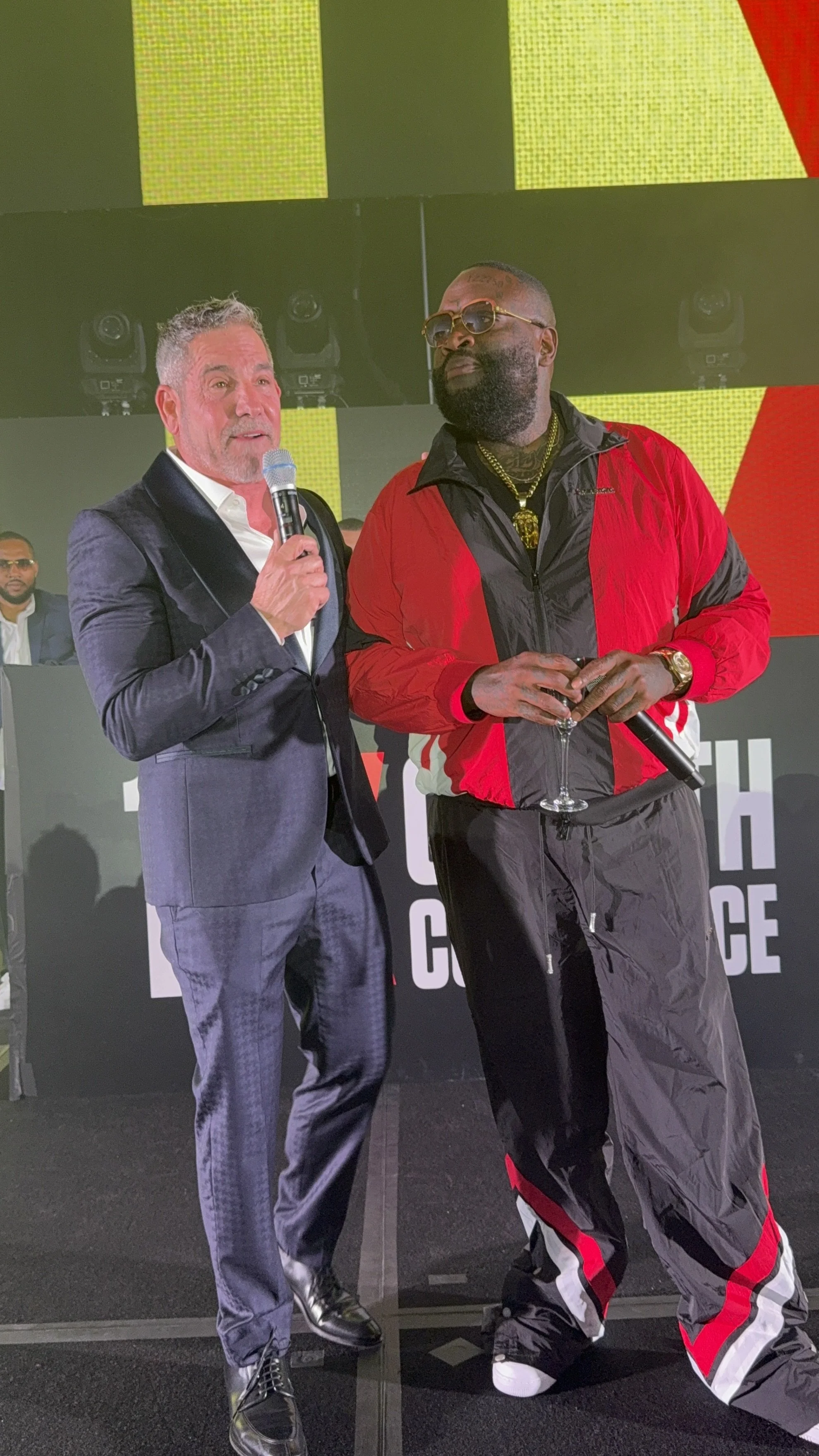 Two men standing on a stage at a conference, one in a dark suit holding a microphone, the other in a red and black tracksuit holding a wine glass, with a DJ and large screen in the background.