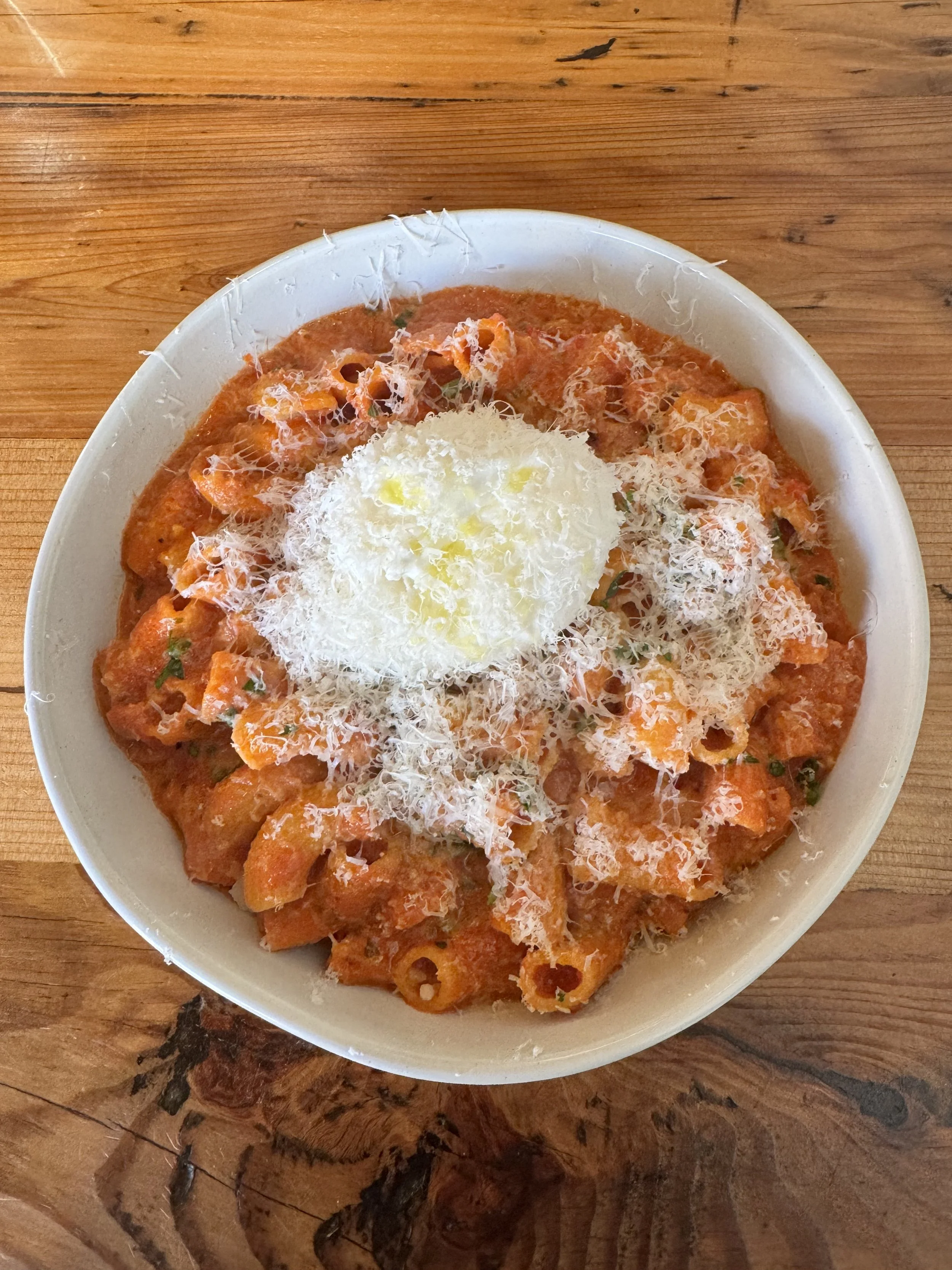A bowl of pasta with tomato sauce topped with shredded cheese and a dollop of ricotta cheese in the center placed on a wooden table.