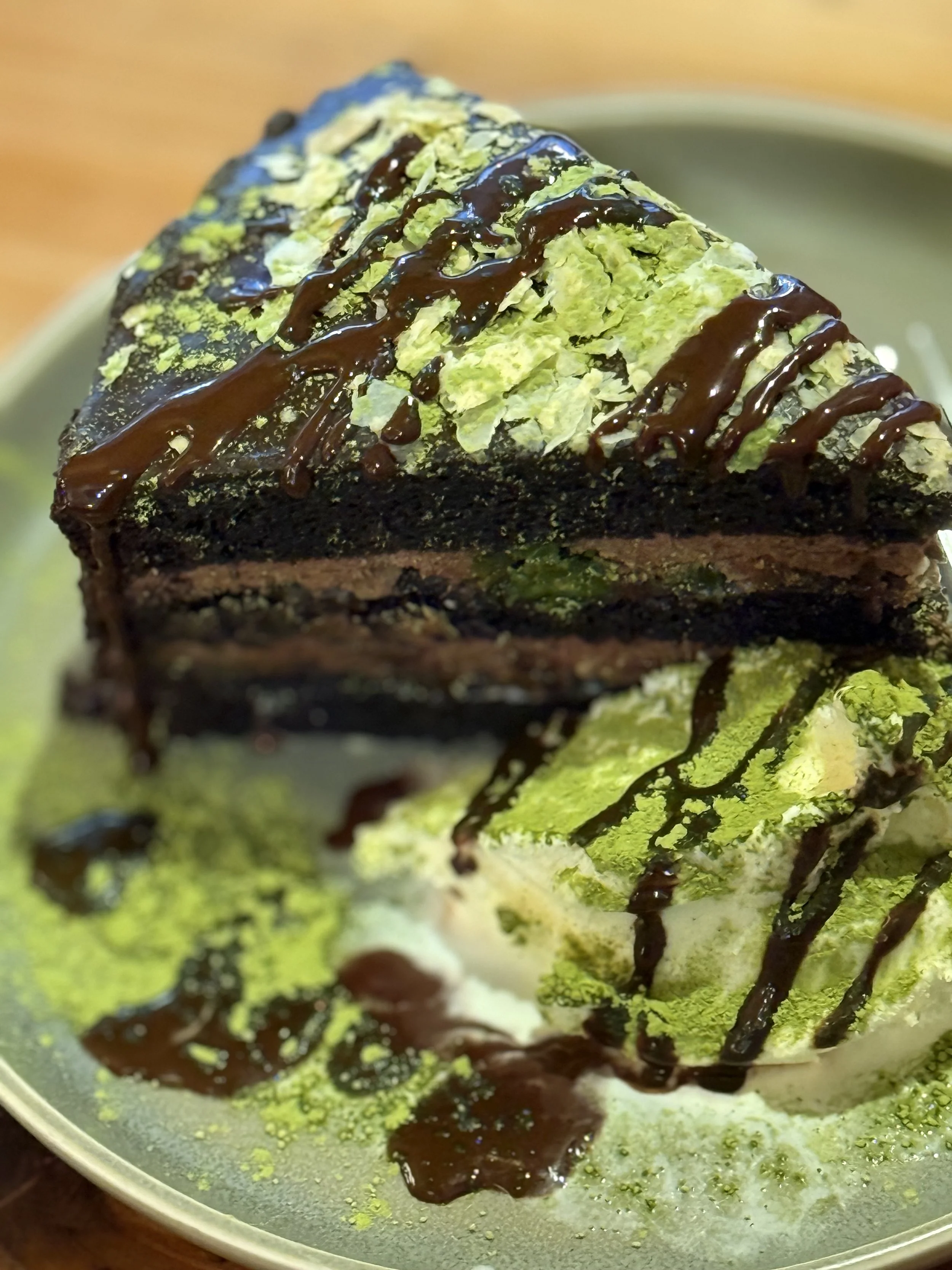 A slice of layered matcha and chocolate cake topped with green powdered matcha, chocolate drizzle, and served with a green matcha-flavored ice cream beside it.