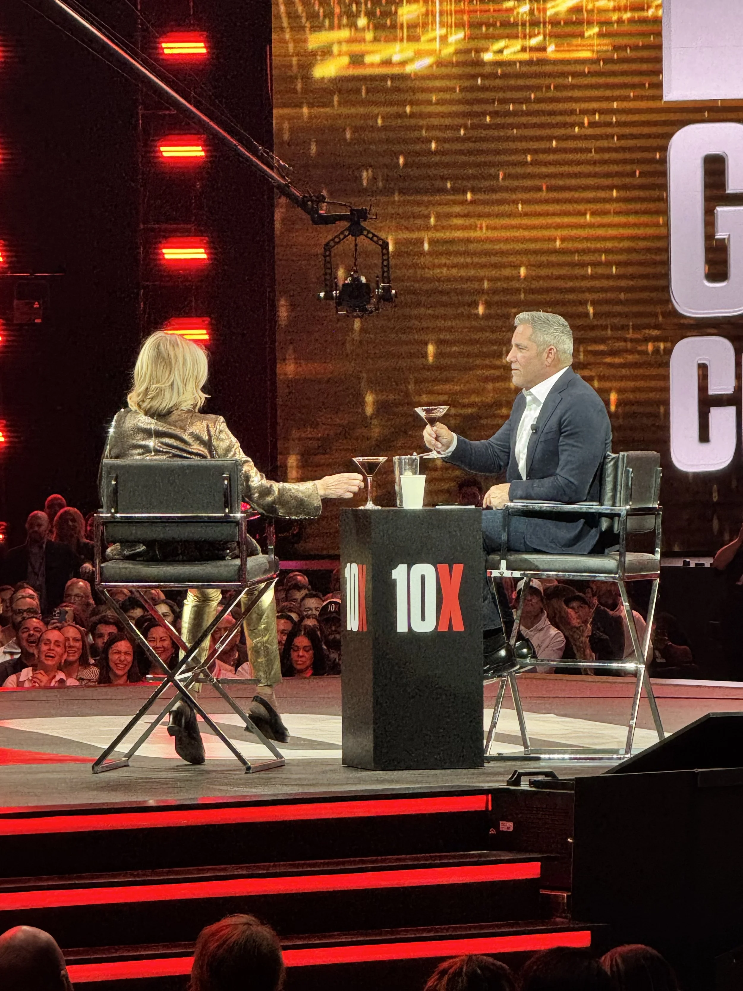 Two people, a woman and a man, sitting on stage in interview chairs, toasting with cocktail glasses. The woman has blonde hair and is wearing a gold jacket, and the man has gray hair and is wearing a dark suit. There is a table with the logo '10X' be