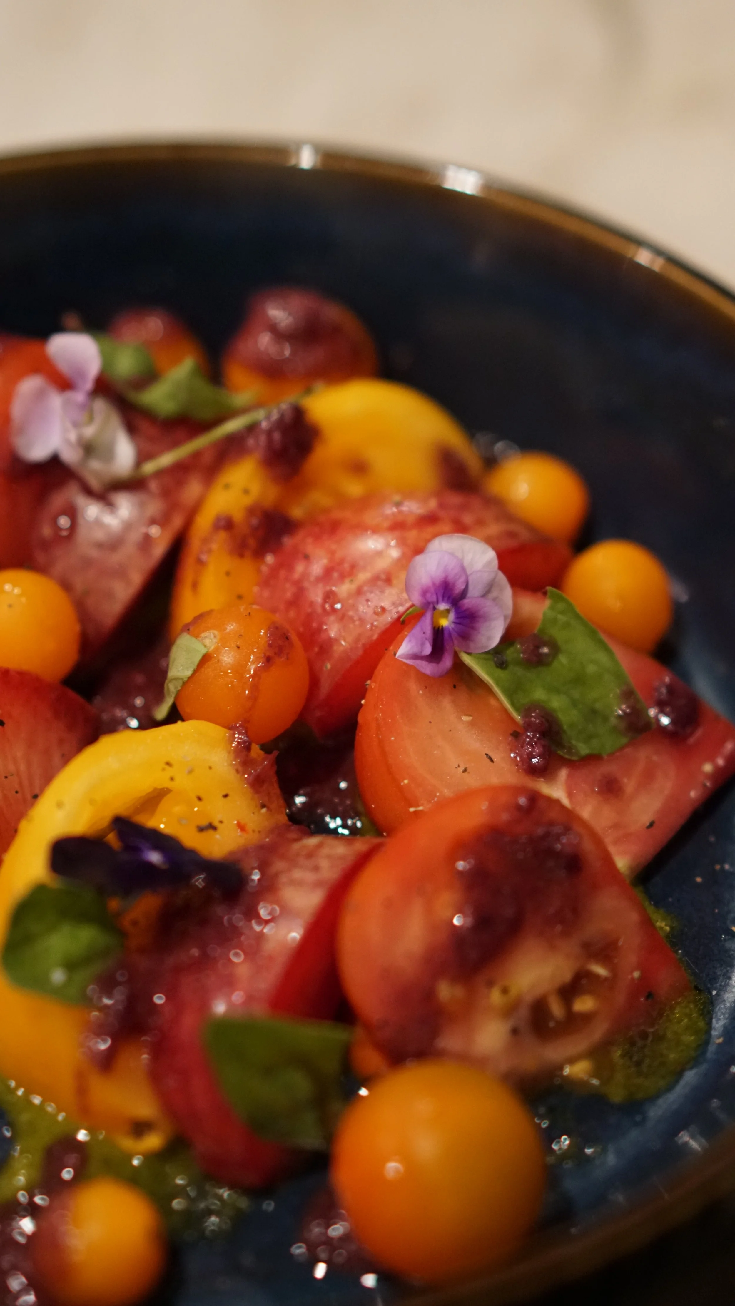 Close-up of a colorful fruit salad with sliced peaches, nectarines, and yellow berries, garnished with edible flowers and small green leaves, served in a dark bowl.