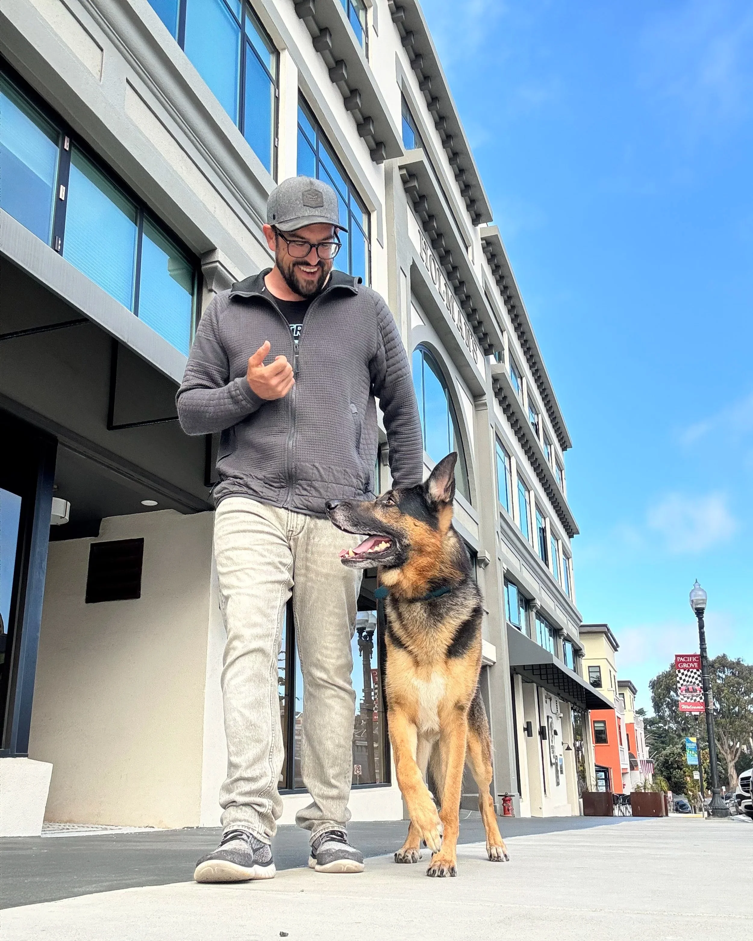 A man smiling and looking at a large German Shepherd dog on a city sidewalk. The man is wearing glasses, a grey cap, a grey zip-up jacket, and light-colored pants. Behind them are modern buildings with large windows and a street sign.