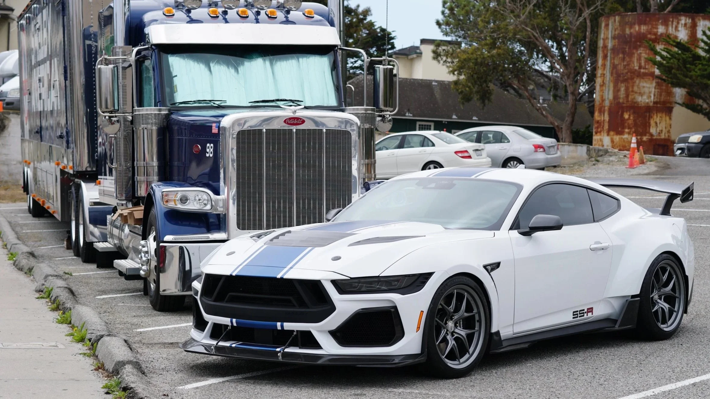 White sports car with black hood and blue racing stripes parked in a parking lot, with a large blue semi-truck behind it.