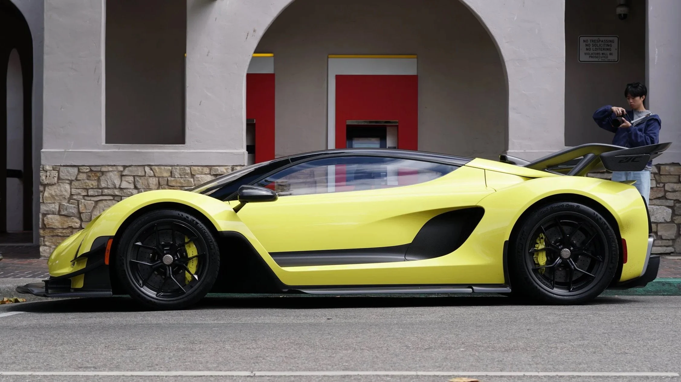 A yellow sports car parked on the street with a person taking a photo in the background.
