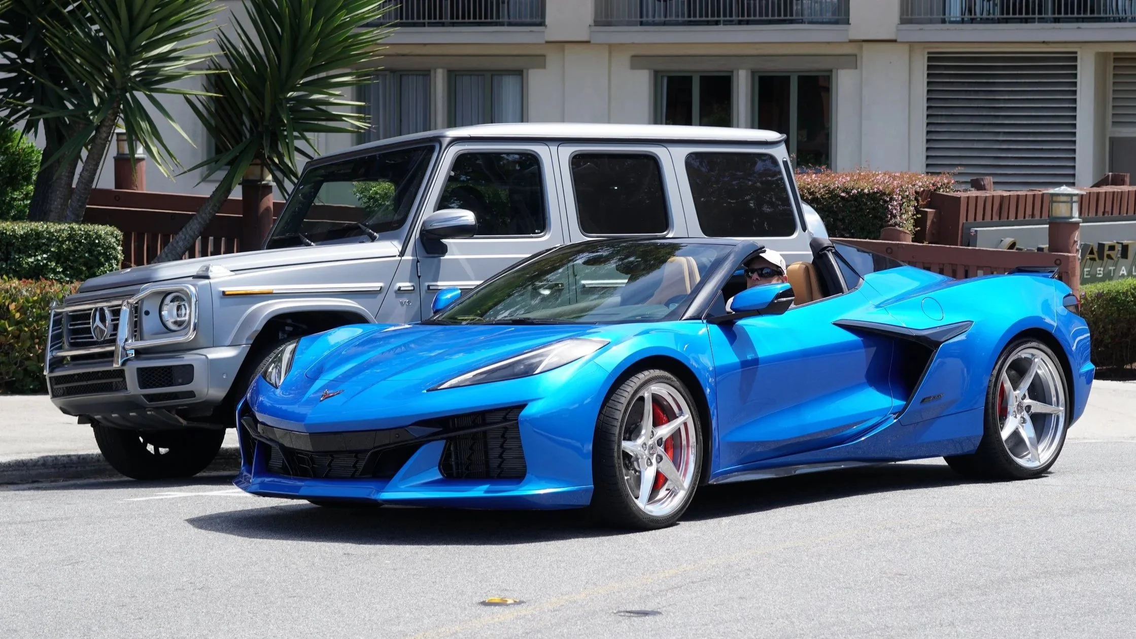 A blue Chevrolet Corvette convertible parked next to a silver Mercedes-Benz G-Class SUV on a city street with residential buildings and greenery in the background.
