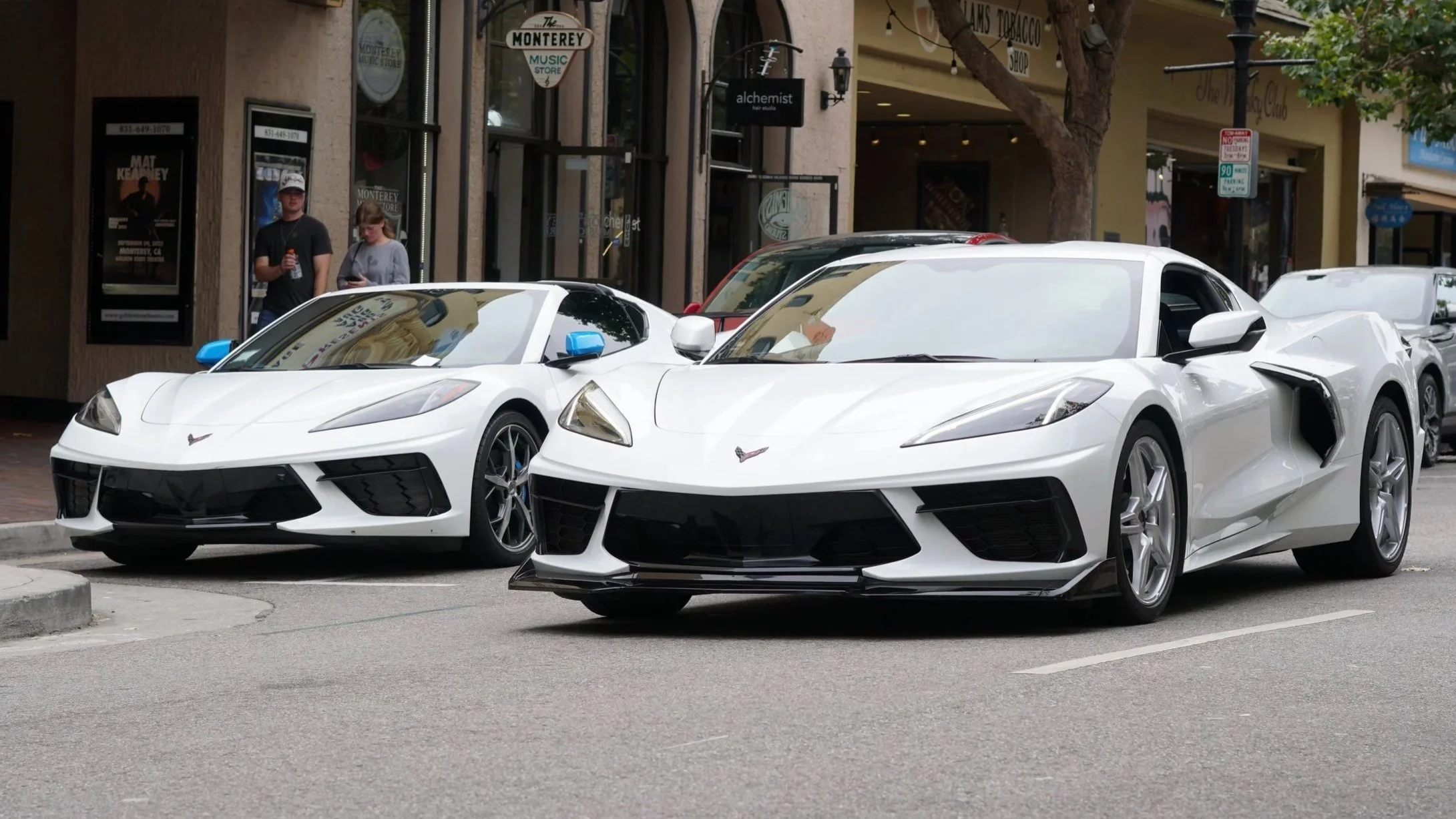 Two white Chevrolet Corvette sports cars parked on a city street, with a storefront and pedestrians in the background.
