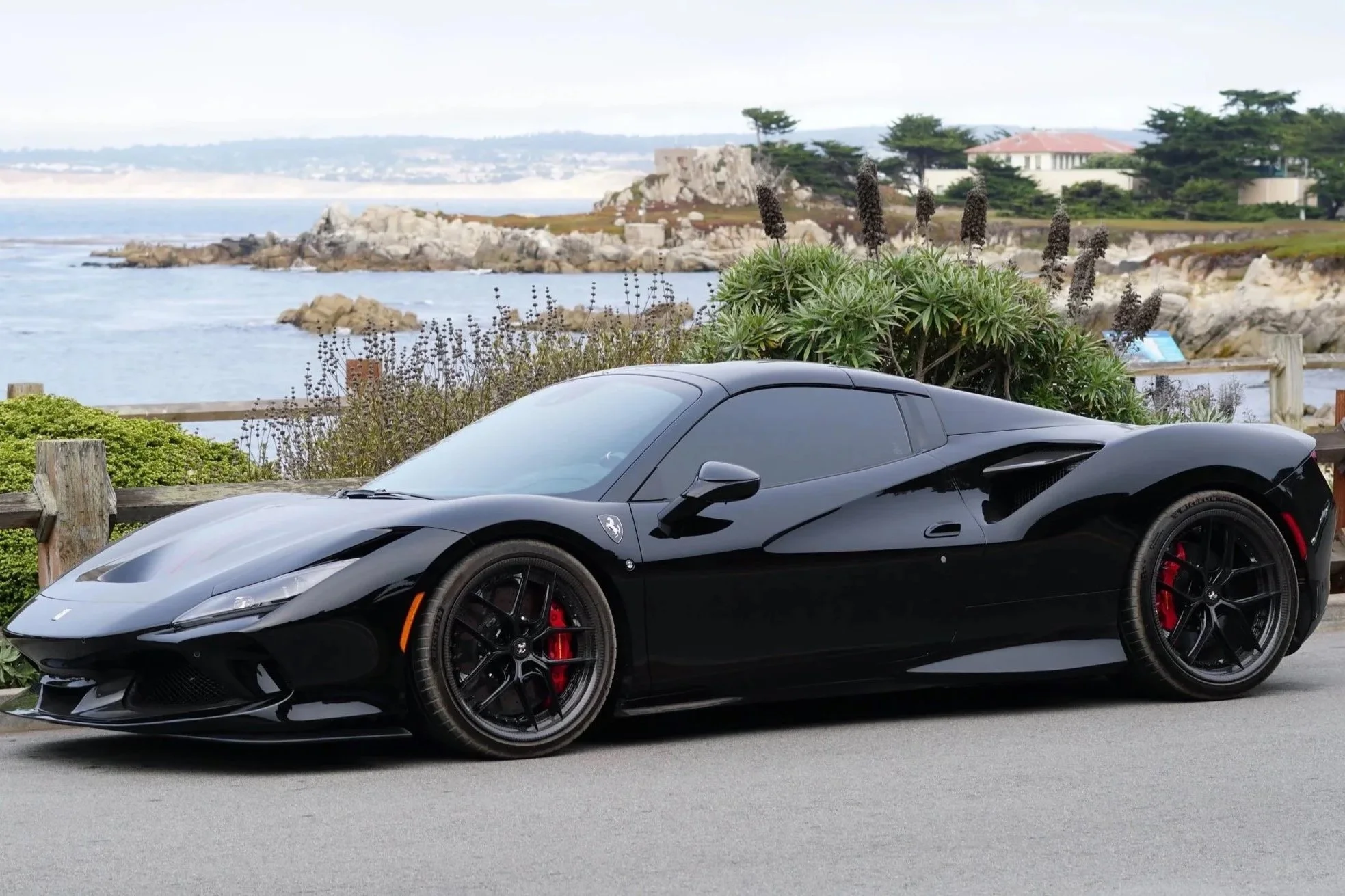 A black Ferrari sports car parked on a coastal road with a view of the ocean, rocky shore, and distant houses in the background.