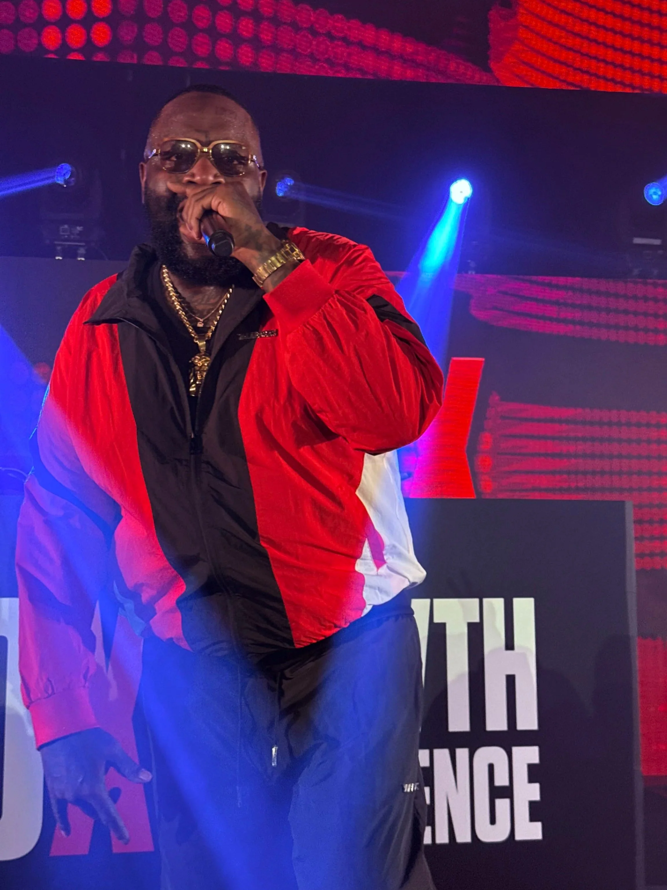 Male performer on stage wearing sunglasses, gold chains, a gold watch, and a red, black, and white jacket, holding a microphone in front of a background with colorful lights and a sign reading 'THE' and 'NCE'.