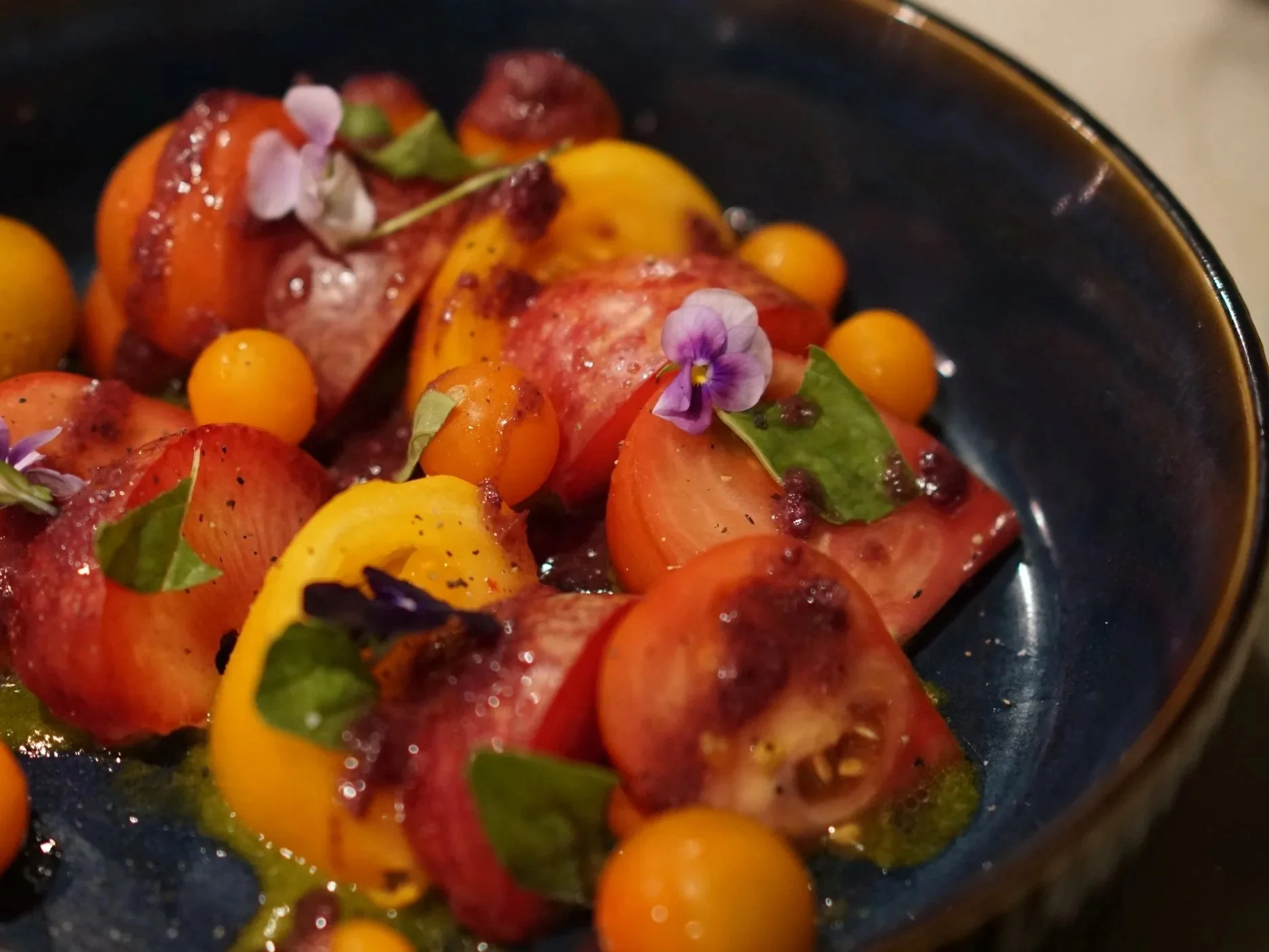 A close-up view of a bowl containing cherry tomatoes, garnished with edible flowers, herbs, and drizzled with oil.