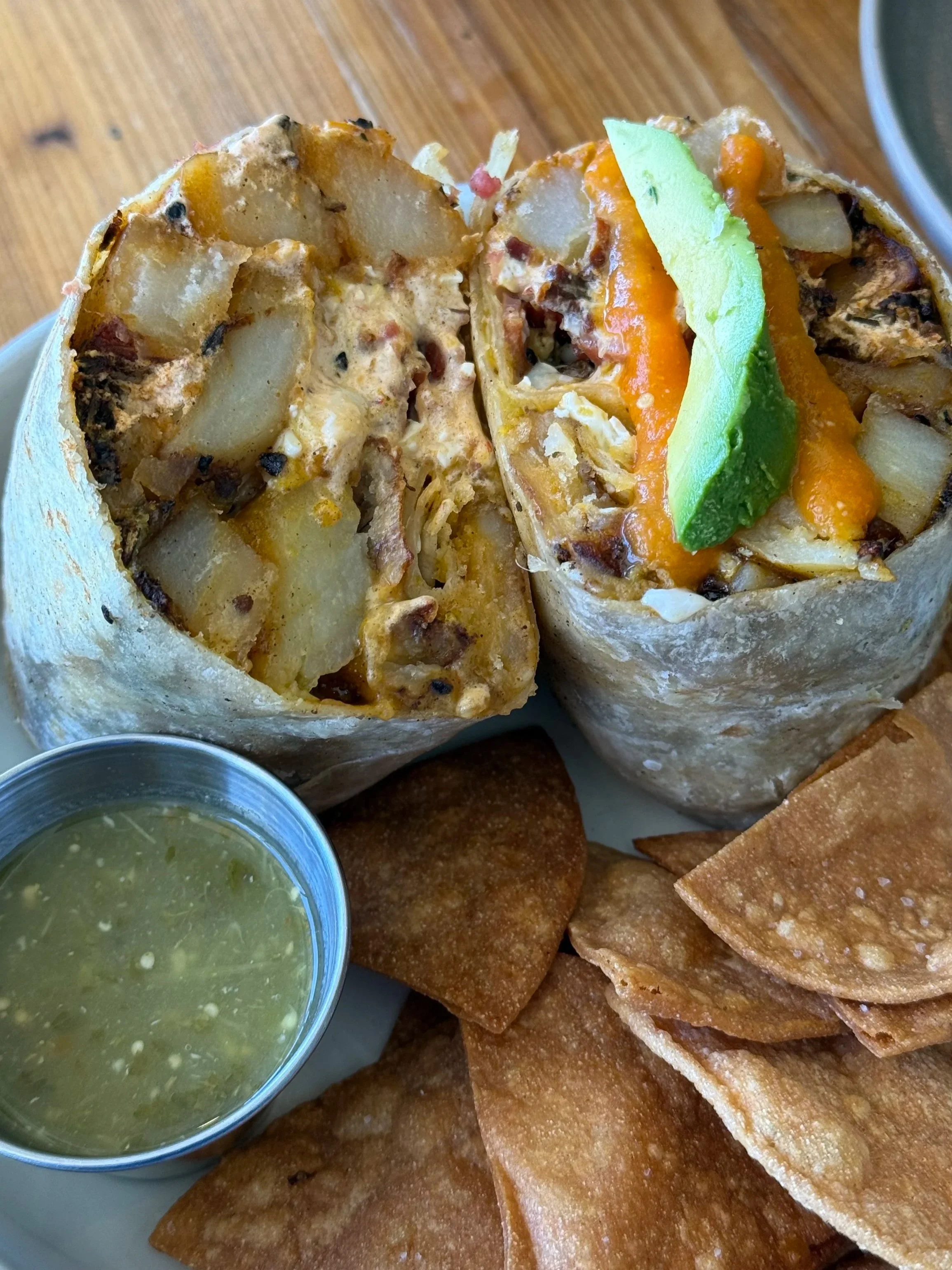 Close-up of a burrito cut in half, filled with potatoes, cheese, and meat, with tortilla on the outside. Next to the burrito are crispy chips and a small cup of green salsa, served on a white plate on a wooden table.