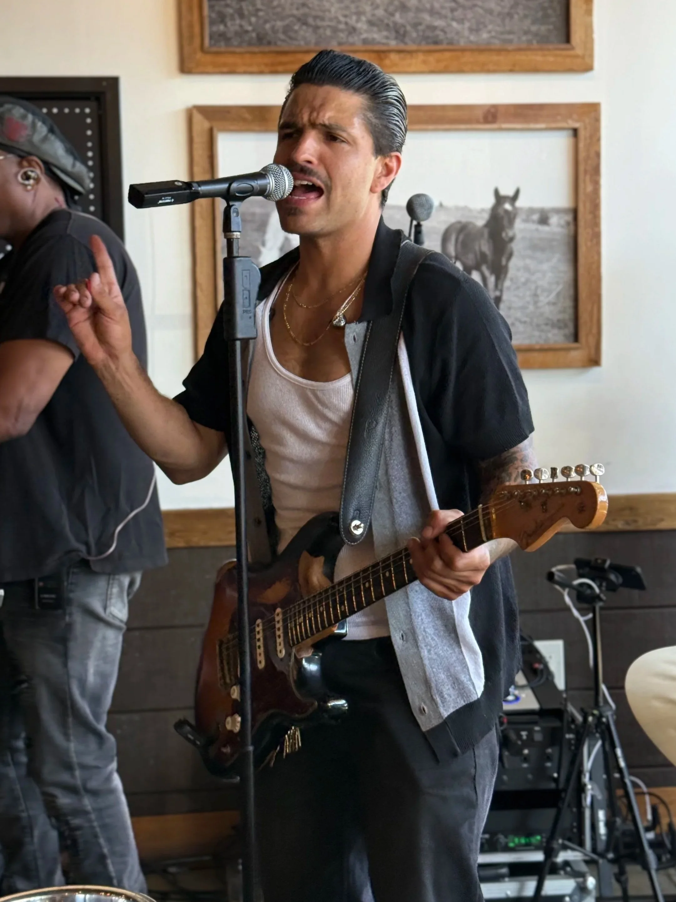 A man singing into a microphone while holding an electric guitar, wearing a black shirt over a white tank top, with slicked-back hair, at an indoor venue with framed black and white photographs on the wall.