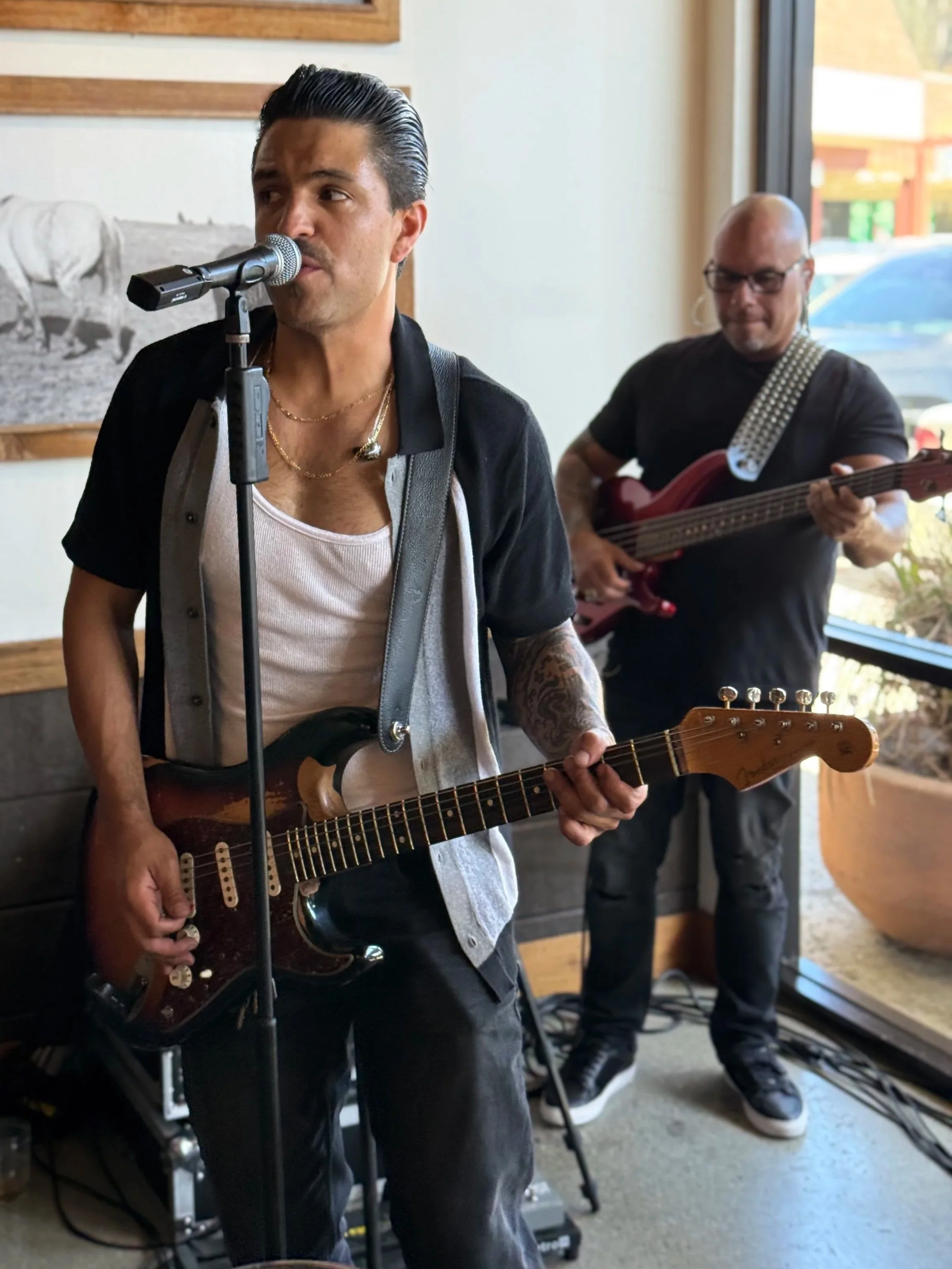 Two men playing musical instruments inside a venue with a large window and a black and white photo on the wall.