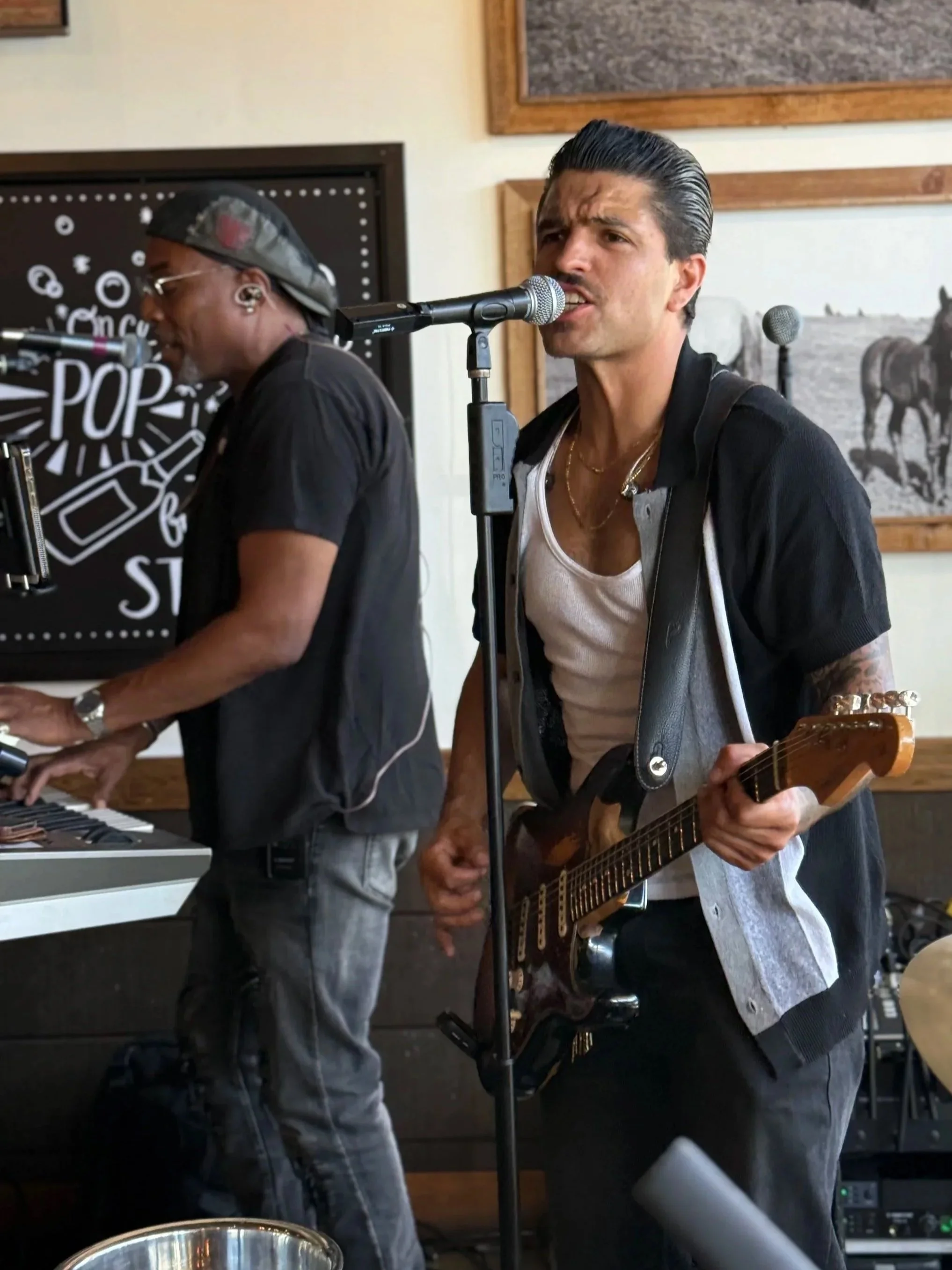 Two musicians performing, one playing keyboard and the other singing and playing guitar in a casual indoor setting.